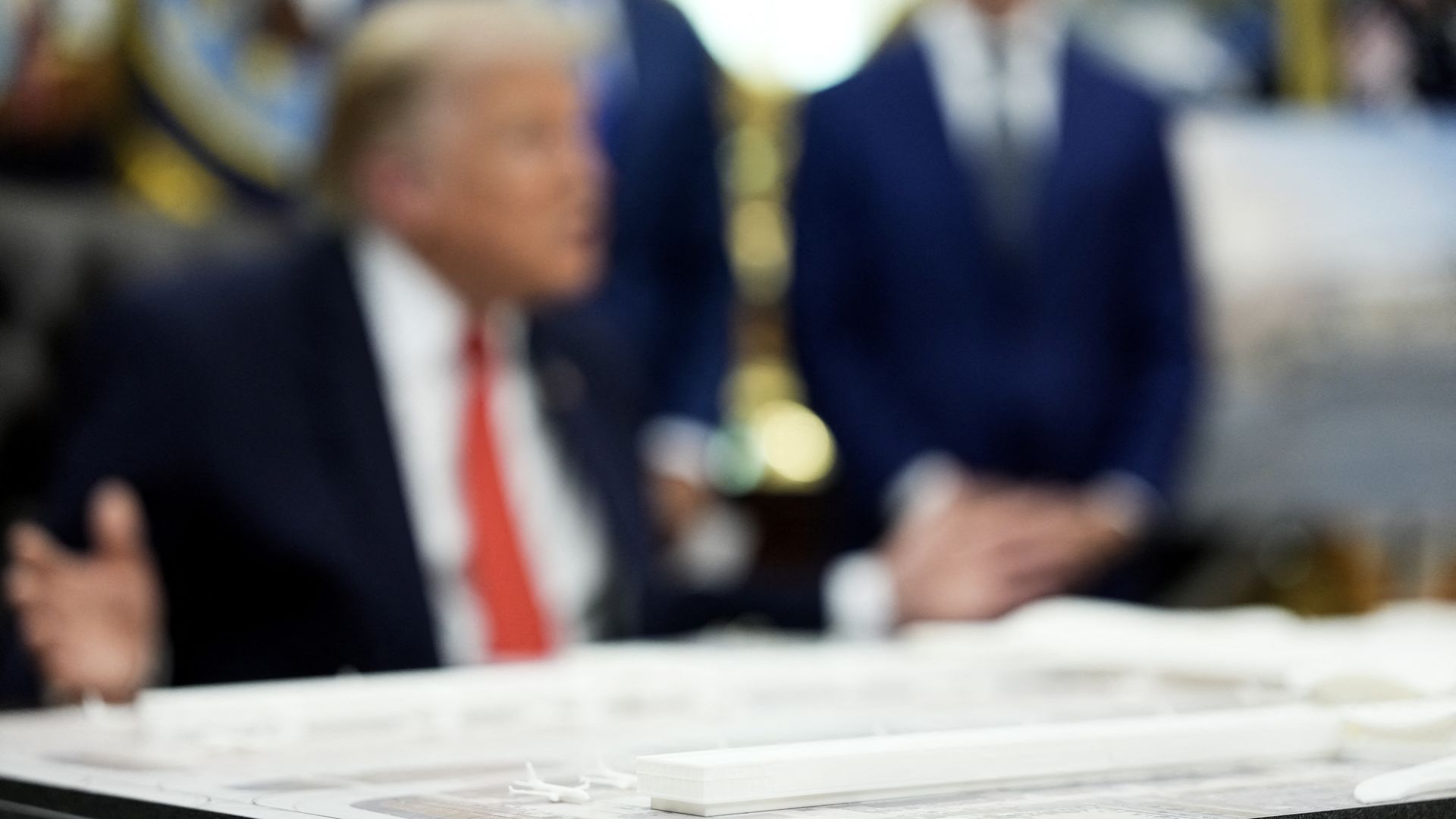 President Trump sits in the Oval Office, out of focus in the picture, in front of a physical mockup of the new Dulles airport