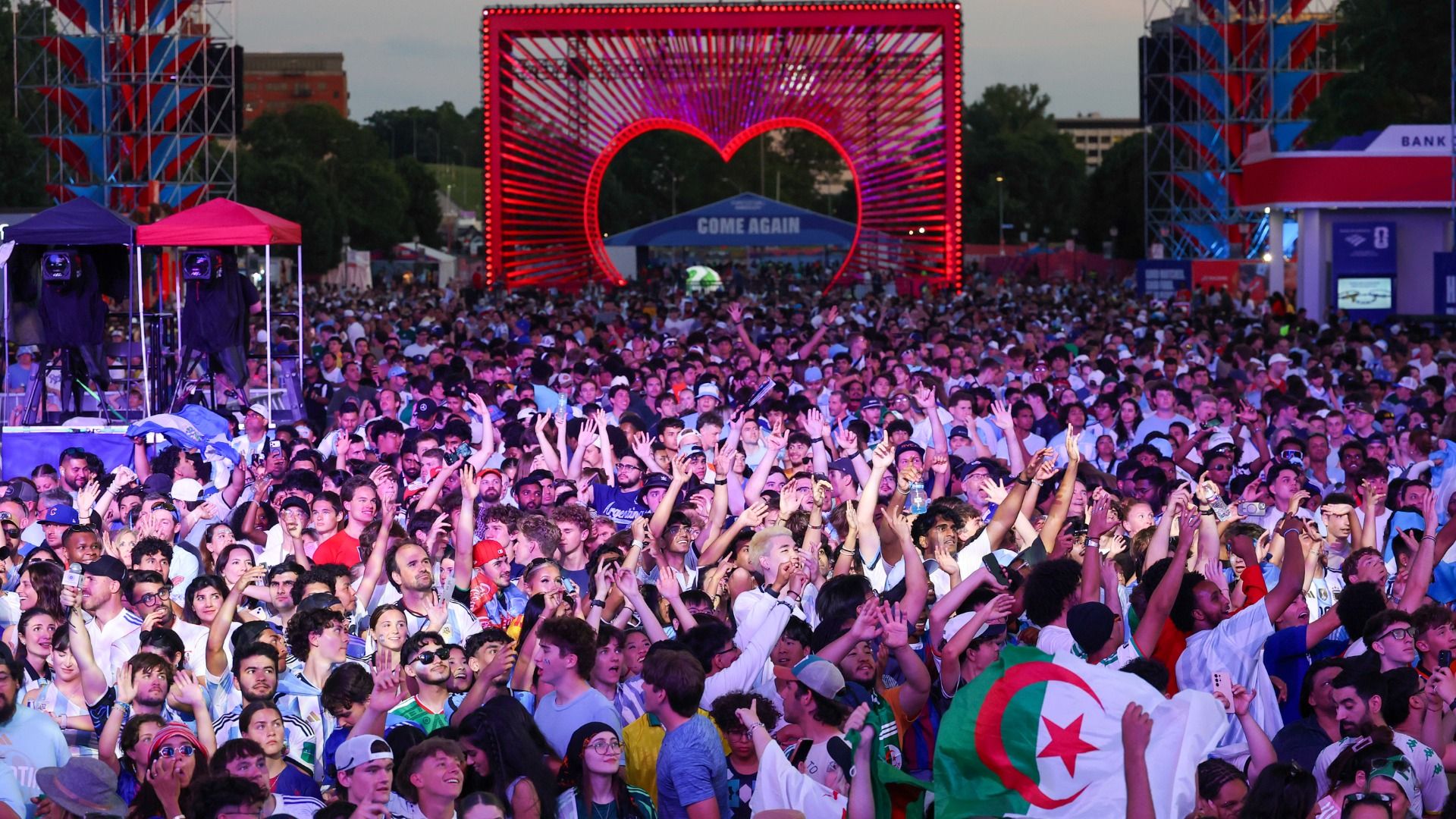 A crowd of fans wearing Argentina and Algeria clothes and flags gather and cheer at Kansas City's Fan Fest, with a big red heart structure in the background and the world "COME AGAIN."