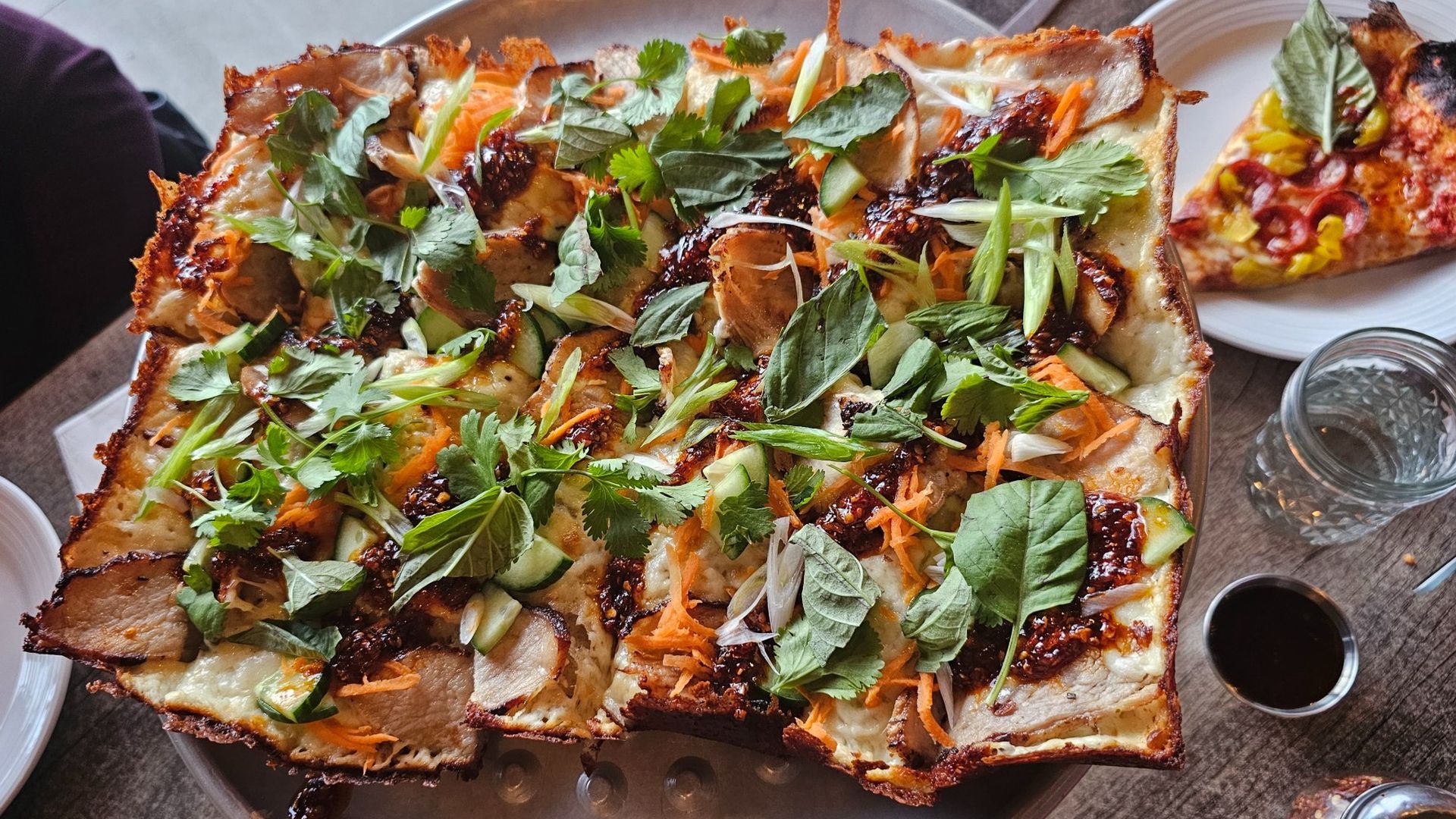Overhead view of a large rectangular flatbread/pizza topped with fresh cilantro, mint, cucumber, shredded carrots and green onions; a dark sauce drips over the crispy crust, served with a side dish and a water glass.