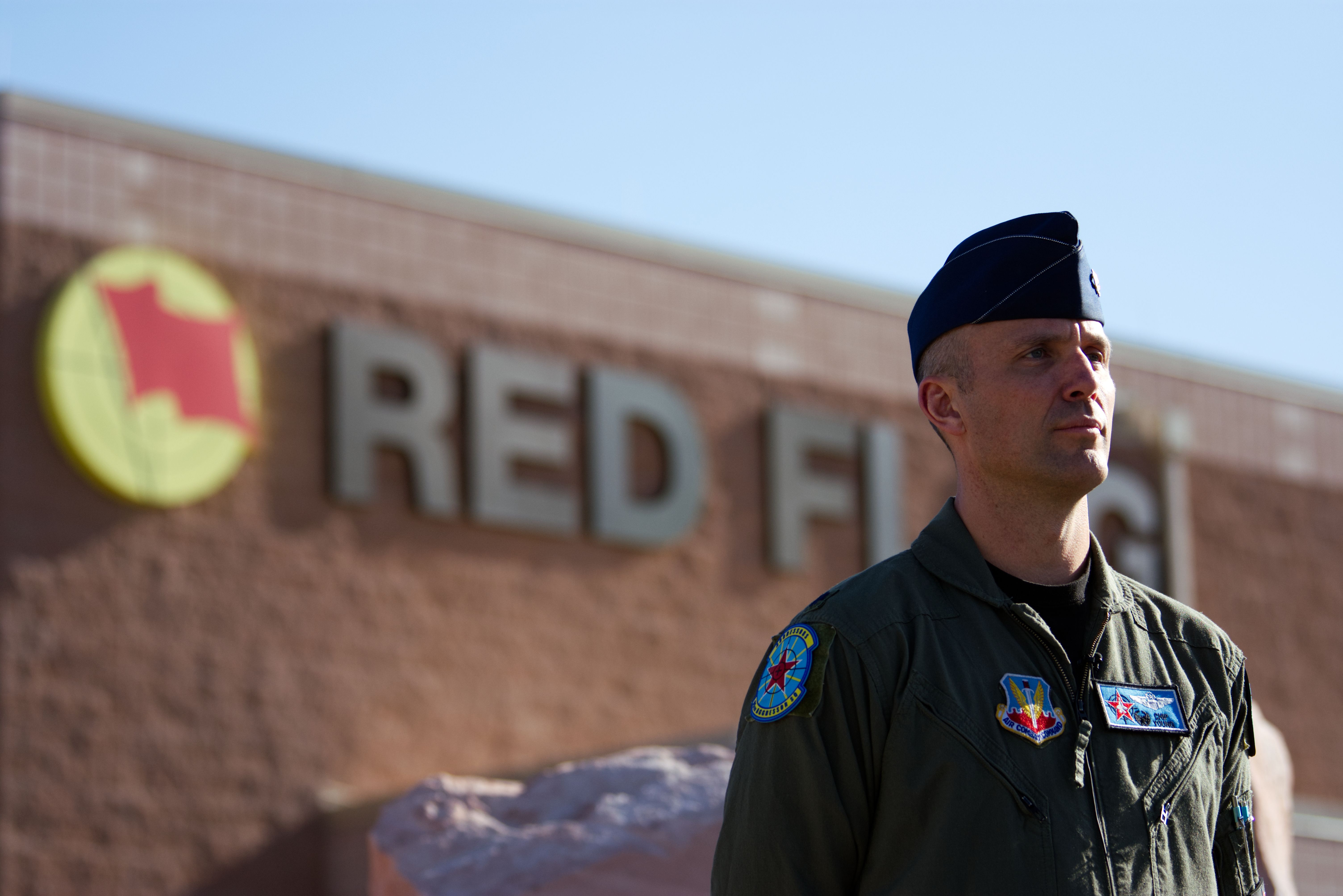 Military man in olive green uniform and blue flight cap stands in front of a building with a blurred sign reading "RED FLAG" and a yellow circle with a red flag symbol.