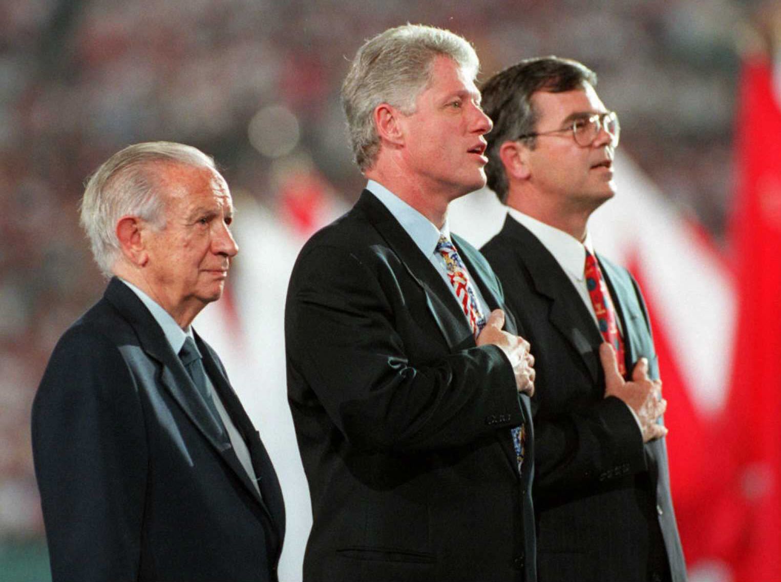 Two individuals wearing suits sing the national anthem with their hands over their hearts while another individual in a suit looks on quietly.