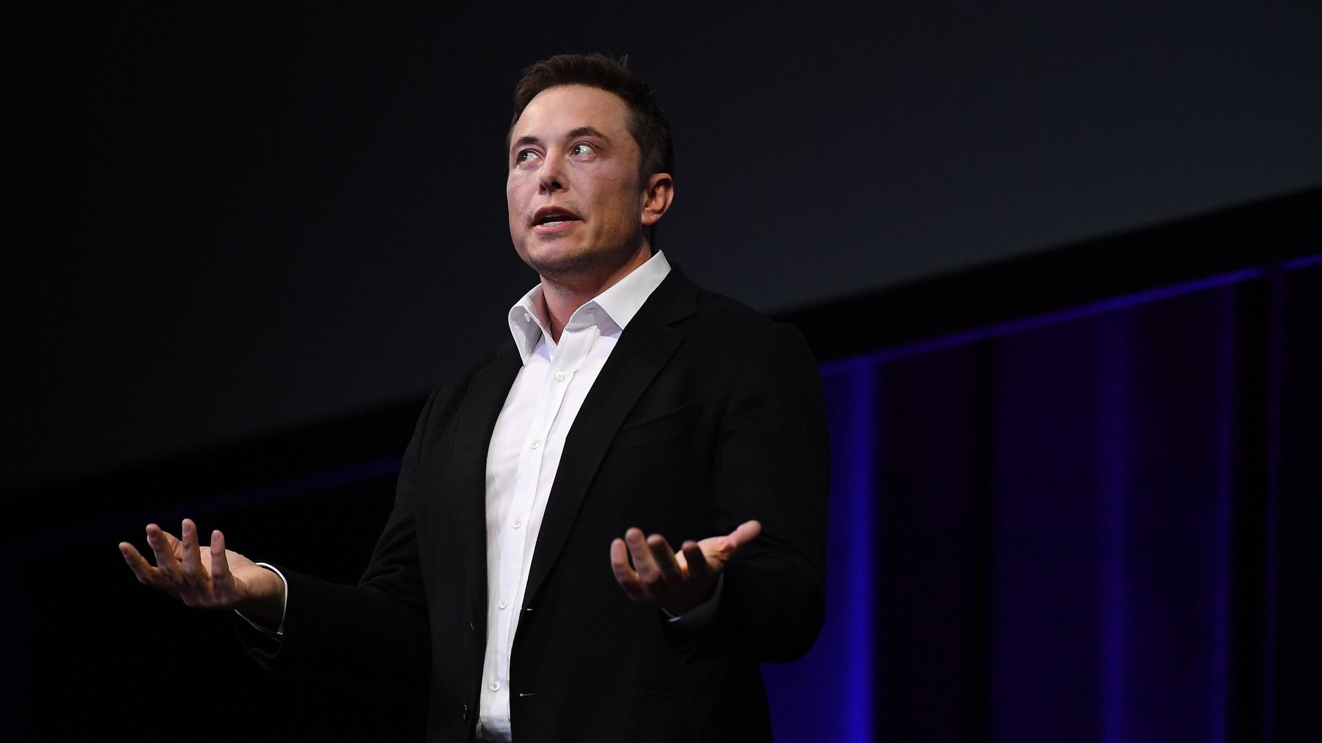Elon Musk in a black suit, white shirt and no tie with both hands palms up talking