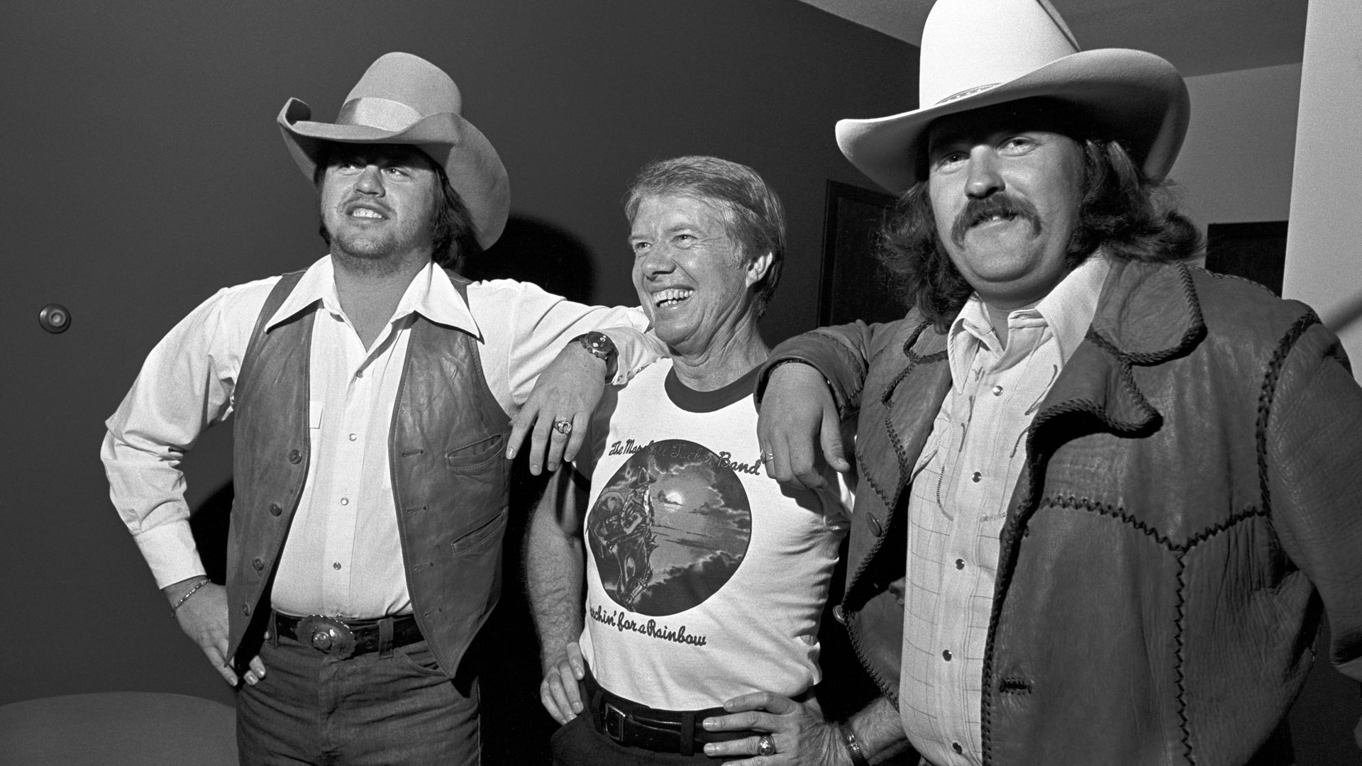 A smiling individual wearing a t-shirt that says "Marshall Tucker Band" stands between two smiling individuals wearing cowboy hats