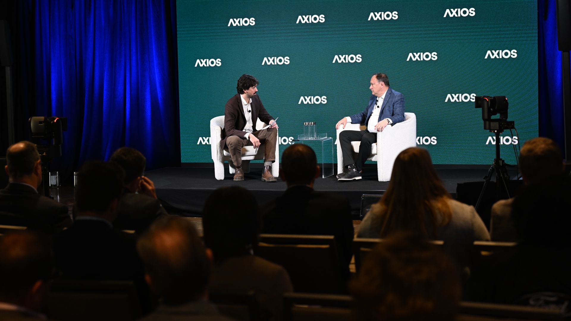 Two men sit on white armchairs on a stage with a teal Axios backdrop. They chat during a panel, with a small glass table between them and an audience in front.