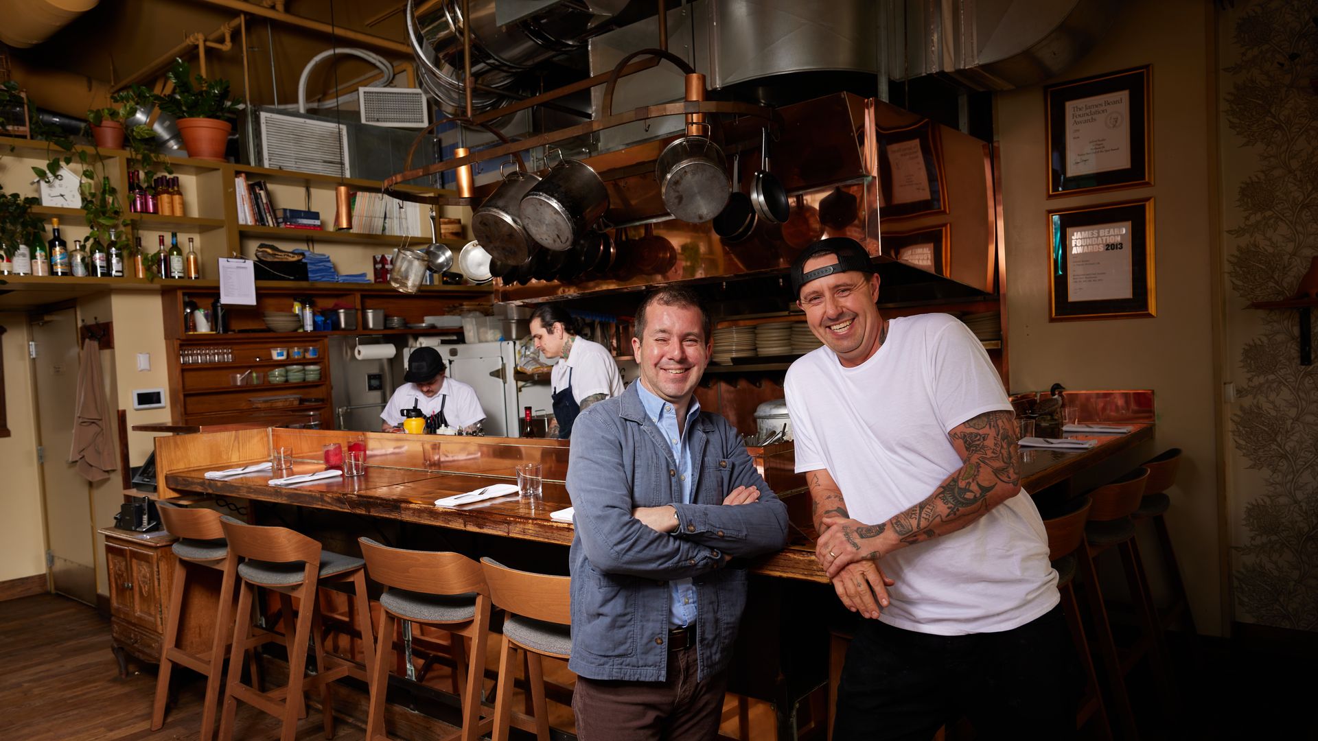 Two people stand facing the camera smiling in front of a counter top restaurant kitchen.