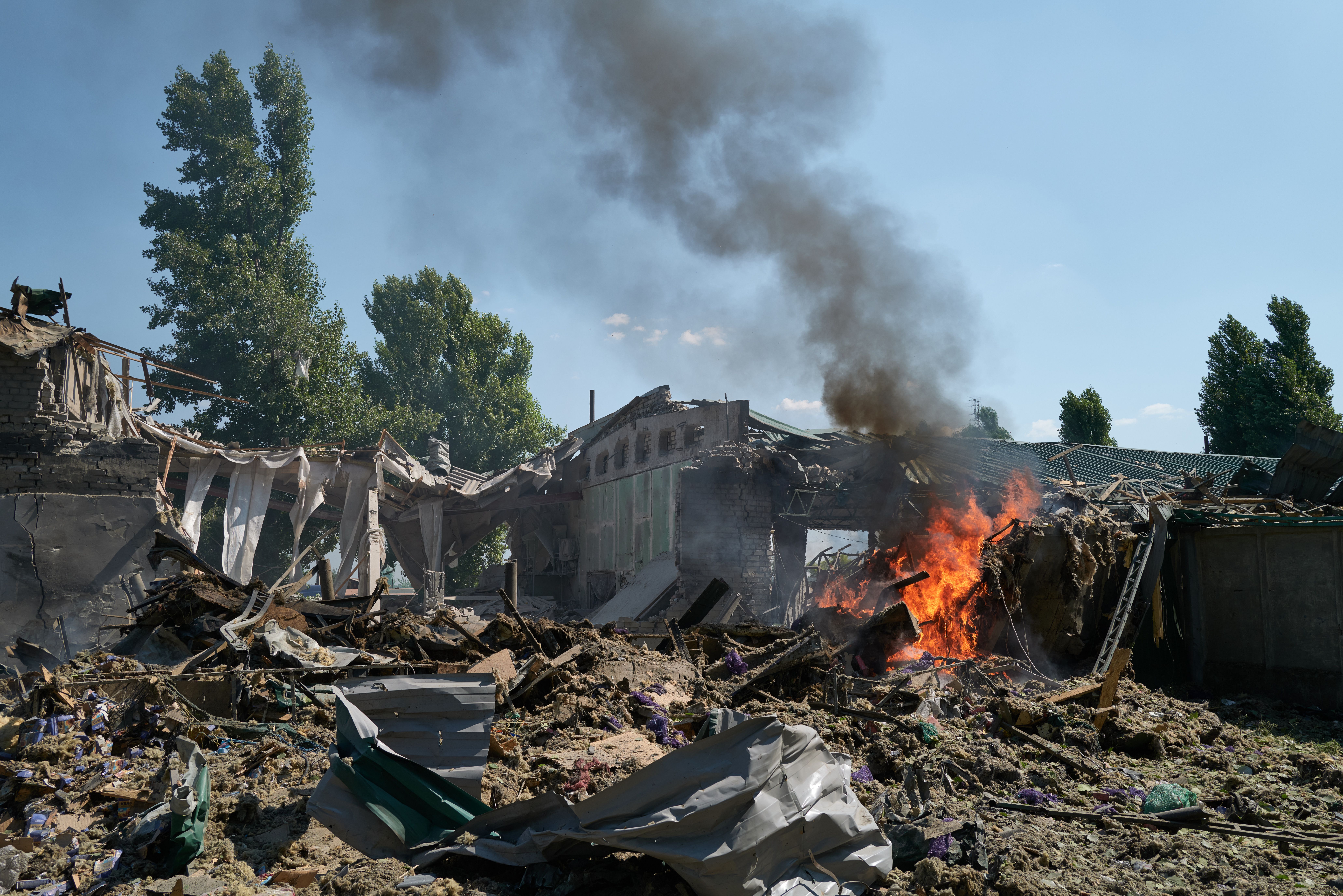 Burning wreckage and debris of a destroyed building with black smoke rising under a clear blue sky, surrounded by tall green trees.