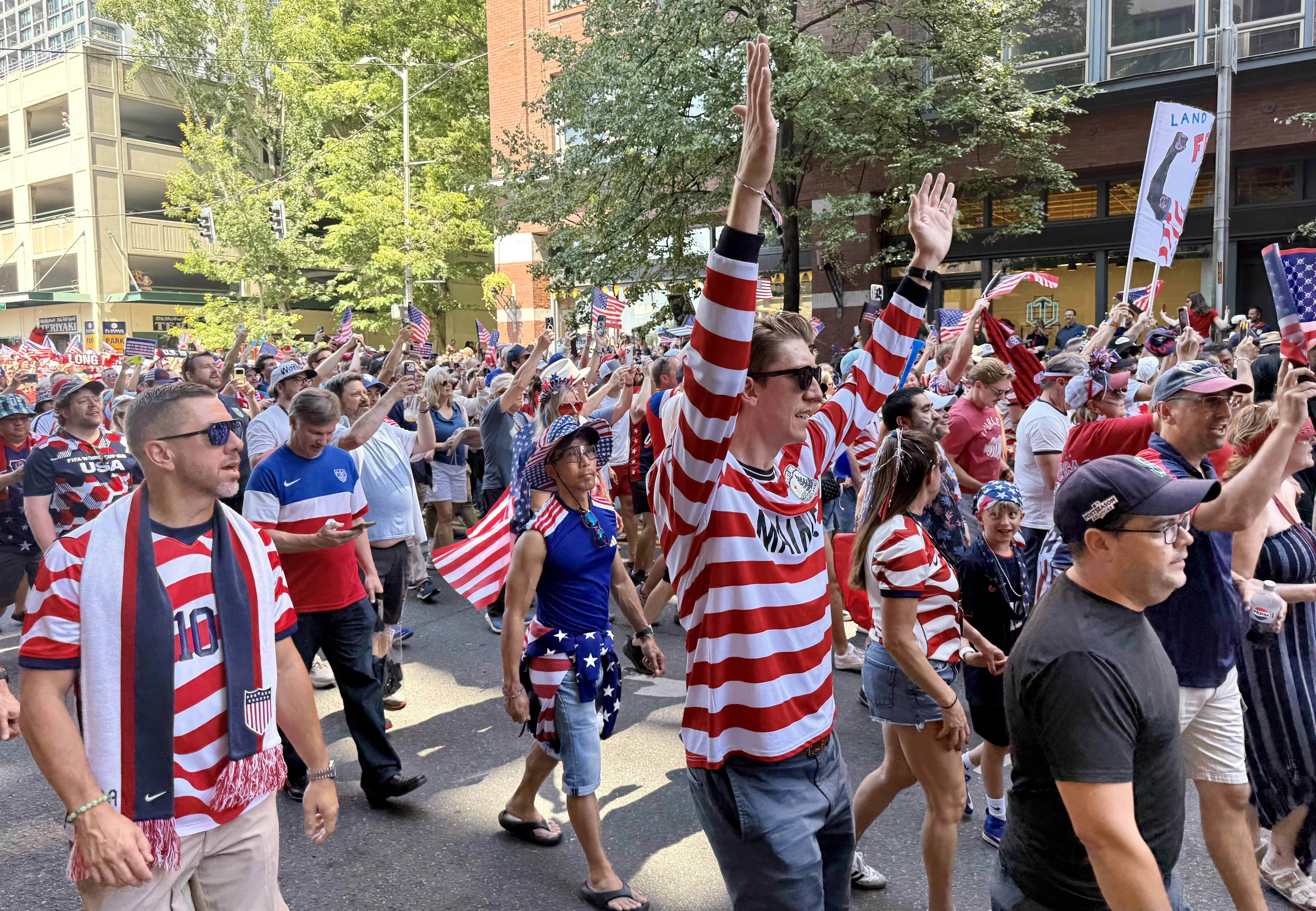 Crowd of parade-goers in red, white, and blue outfits waving small American flags along a city street, with trees and tall buildings in the background.