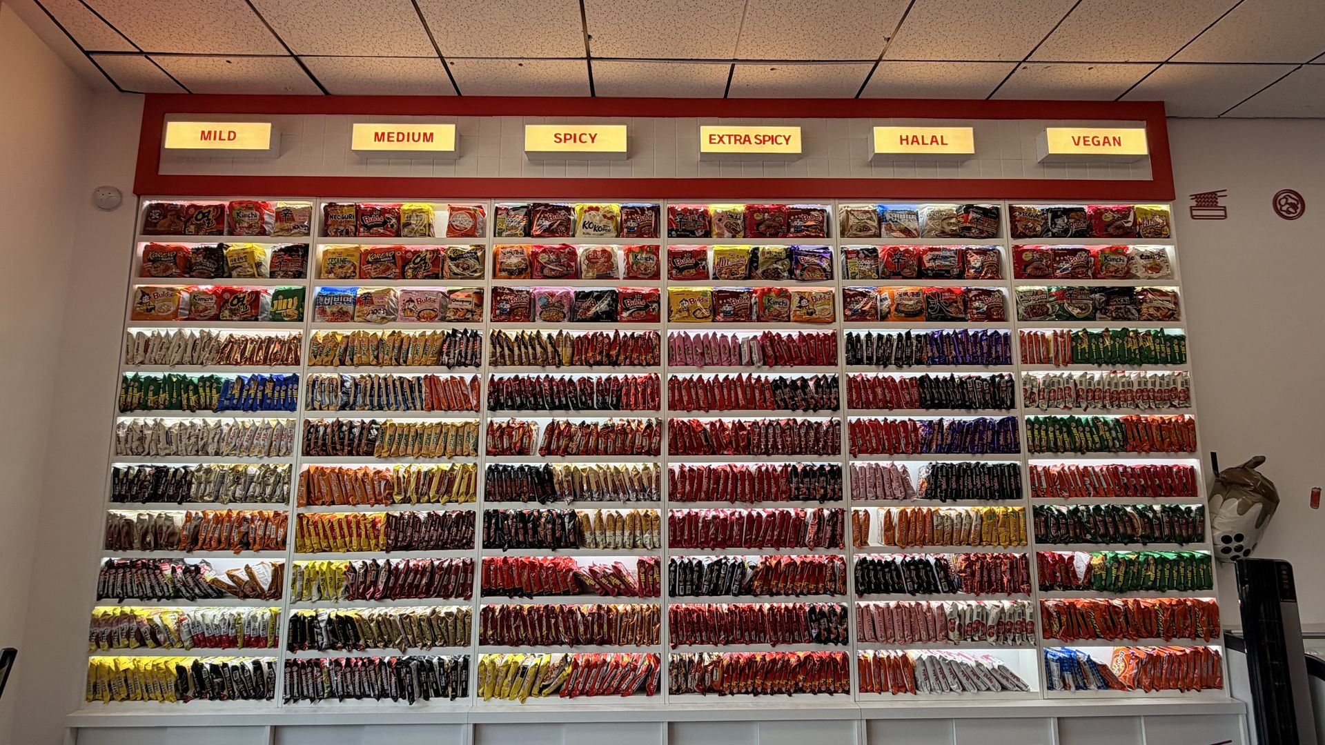 Colorful snack wall with rows of chip bags on white shelves; overhead signs read MILD, MEDIUM, SPICY, EXTRA SPICY, HALAL, VEGAN, indicating spice levels and dietary options.