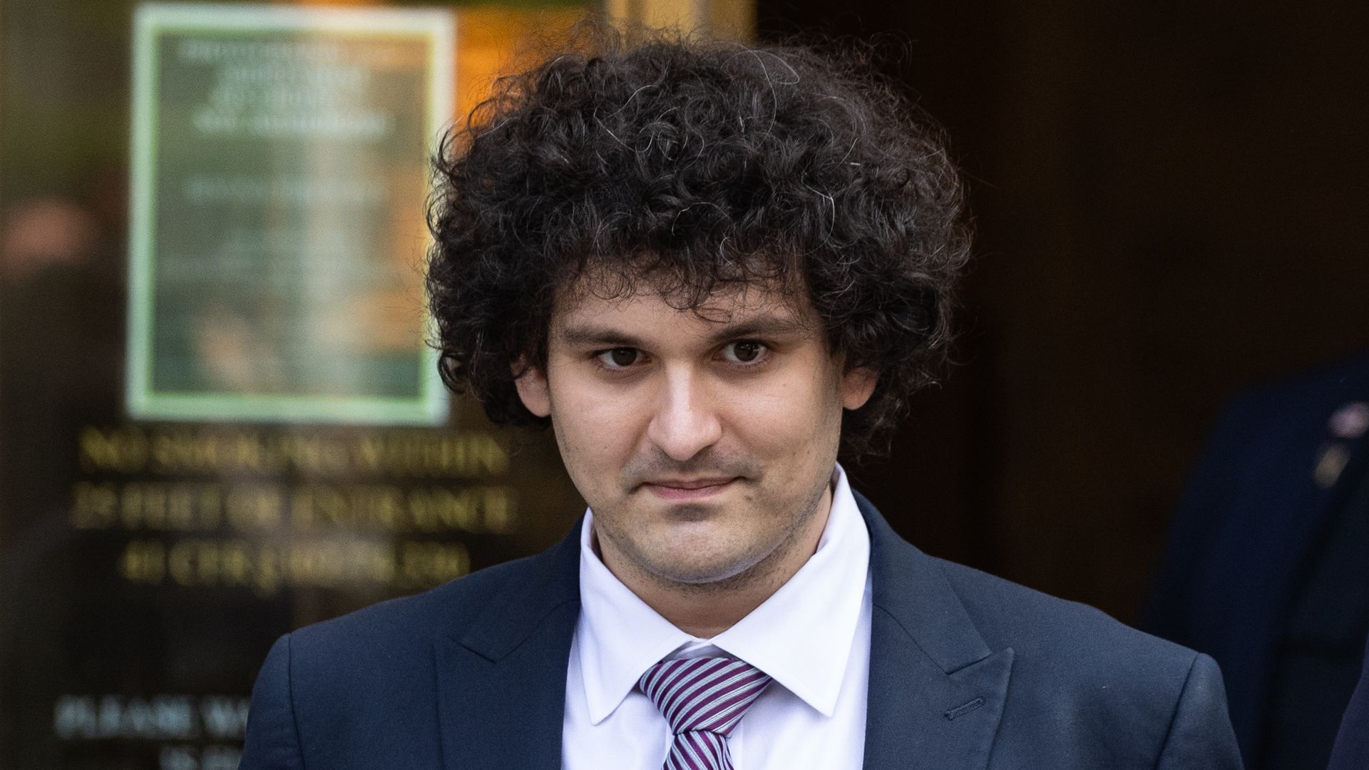 Man with dark, curly hair in a navy suit and striped tie, looking at the camera with a subtle smile; blurred background board behind him.