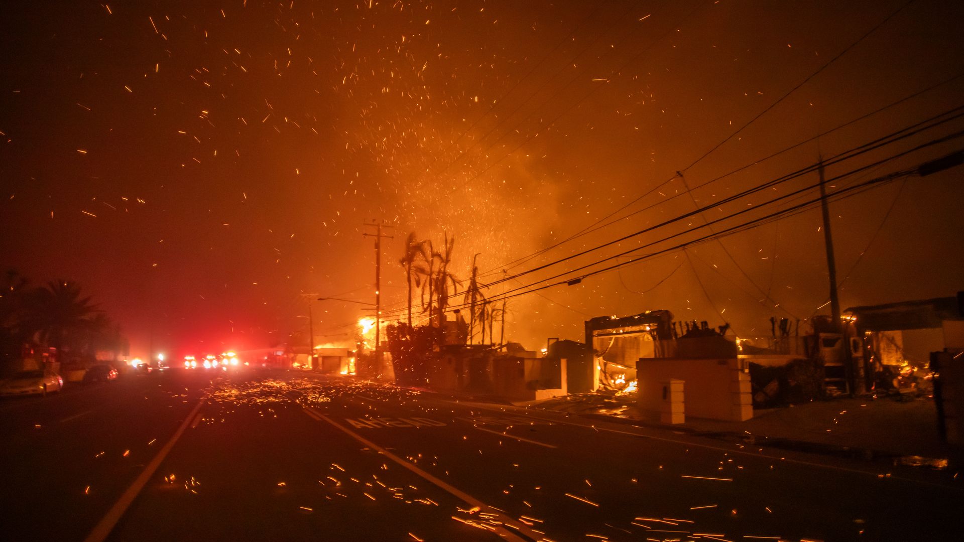 Burning embers fill the air along a street lined with burned buildings in the dark, with emergency vehicles in the background.