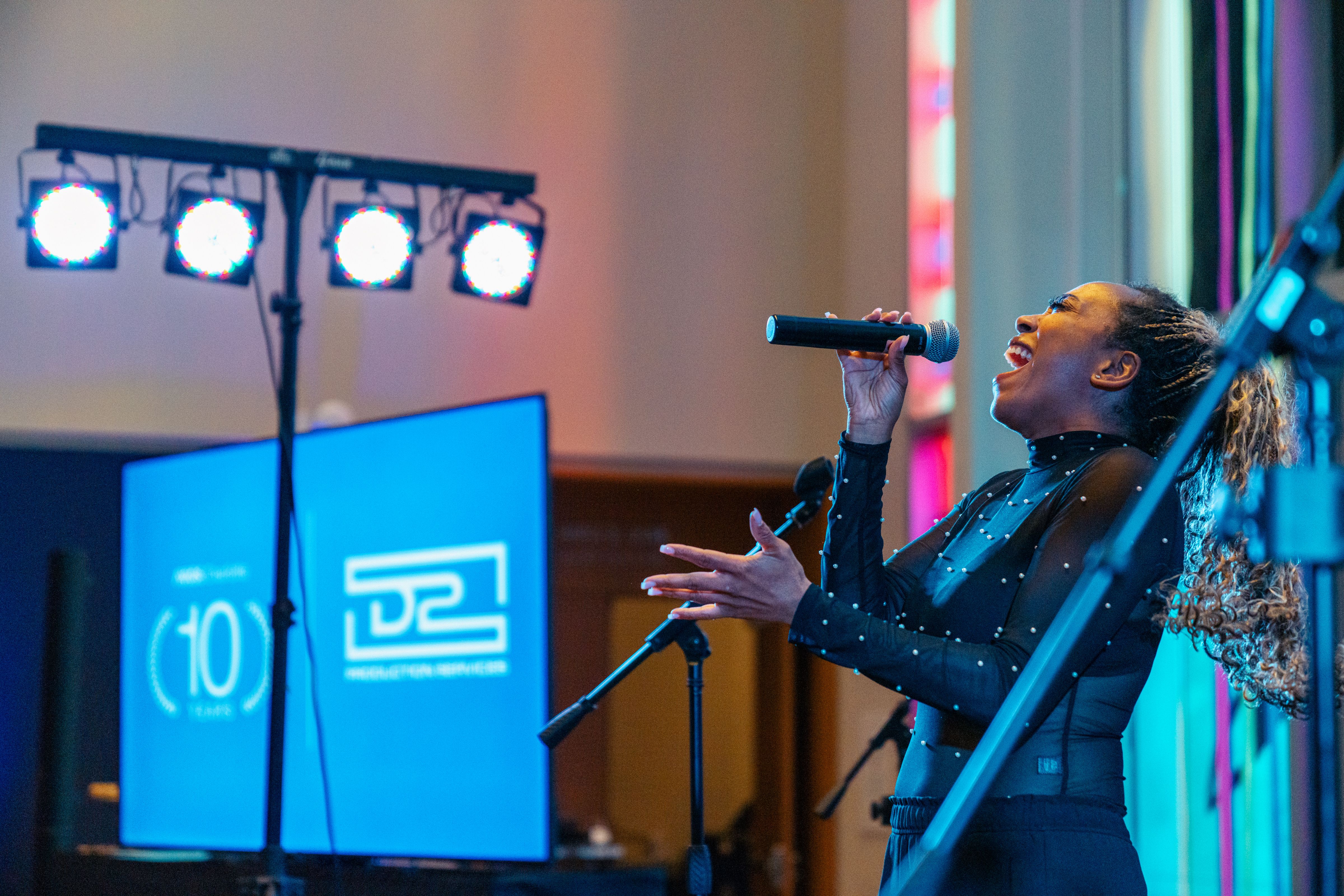 Woman with curly hair in a black sheer long-sleeve top singing passionately into a microphone on stage with blue-lit screens and bright stage lights behind her.