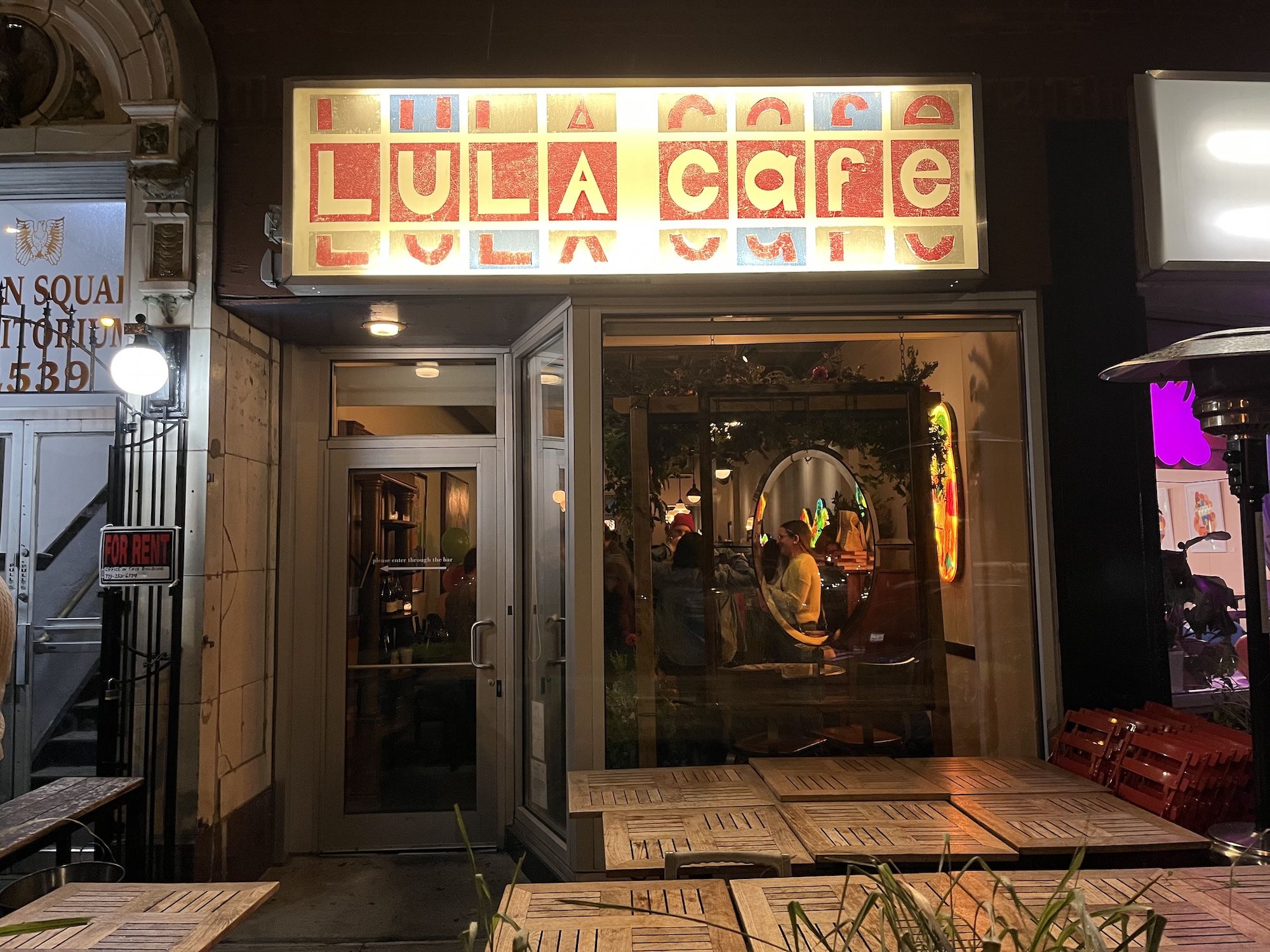 Night view of Lula Cafe with illuminated red and white sign, glass door with text, wooden outdoor tables, and people visible inside through large window with decorations.