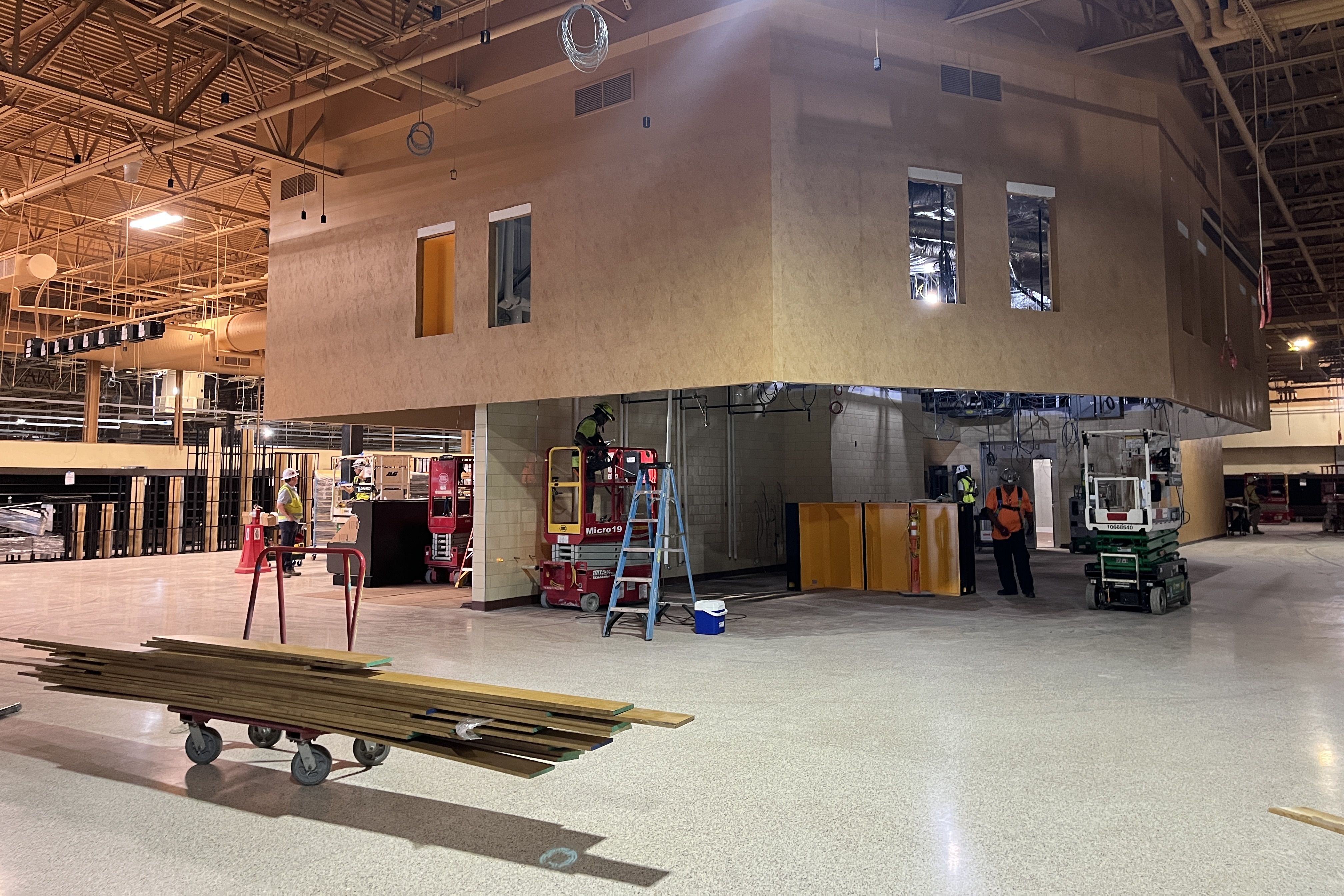 Indoor construction site with a large beige suspended box structure featuring three window openings. Workers in hard hats, ladders, a red scissor lift, and stacks of lumber under an exposed ceiling with pipes.