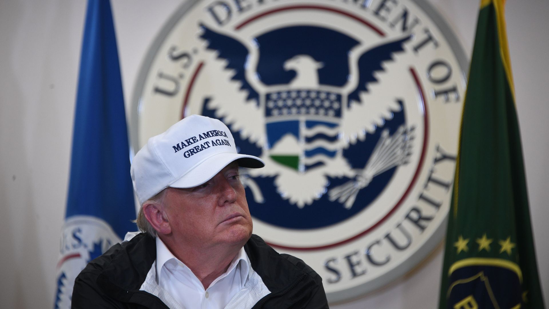 Trump sits with a white MAGA hat on in front of the logo for the US Department of Homeland Security 