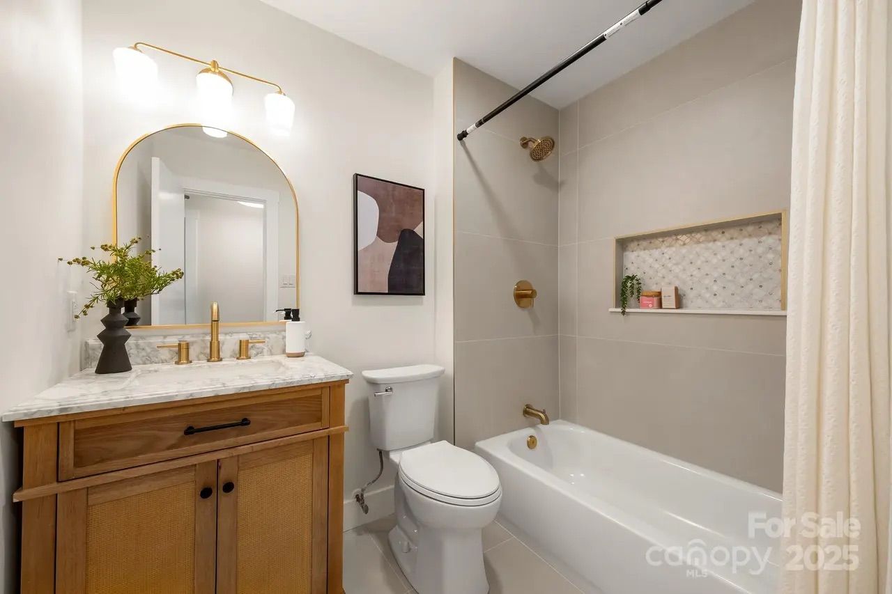 Modern bathroom with wooden vanity, marble countertop, gold fixtures, white toilet, bathtub with beige tiles, a small plant, and abstract wall art. Soft lighting above large mirror.