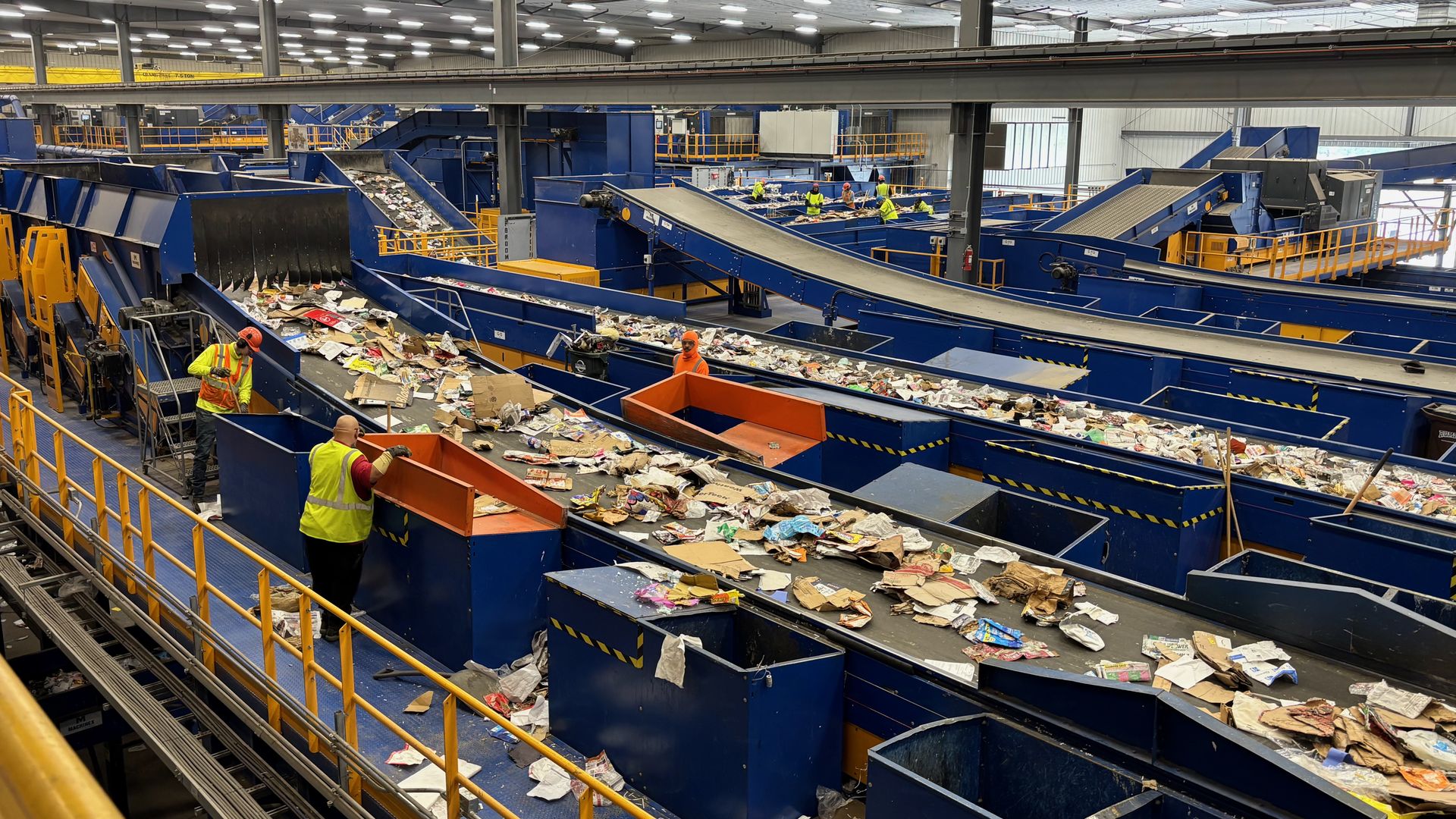 Industrial recycling facility with blue conveyors and sorting bins; workers in yellow safety vests monitor moving waste amid piles of paper and cardboard under bright overhead lights and orange railings.
