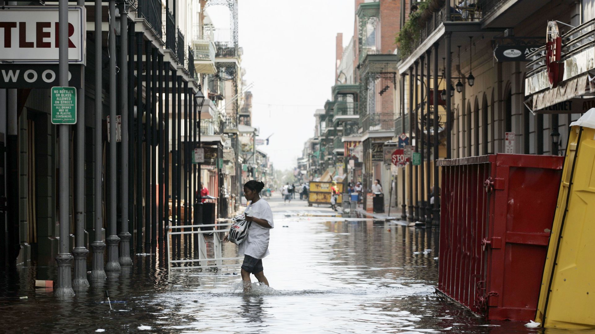 Person walking through a flooded urban street with water reaching mid-calf, surrounded by buildings with balconies, a red container, and yellow portable toilet in the water.