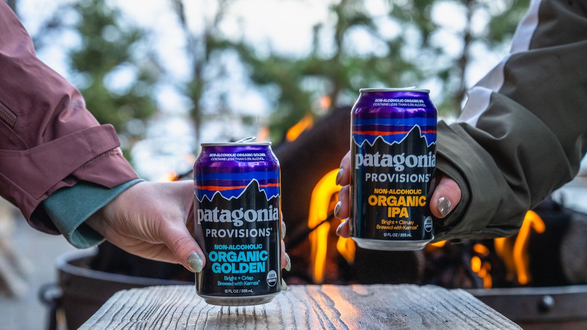Two hands holding cans of Patagonia Provisions non-alcoholic organic beer, one labeled "Organic Golden" and the other "Organic IPA," near a fire pit outdoors.