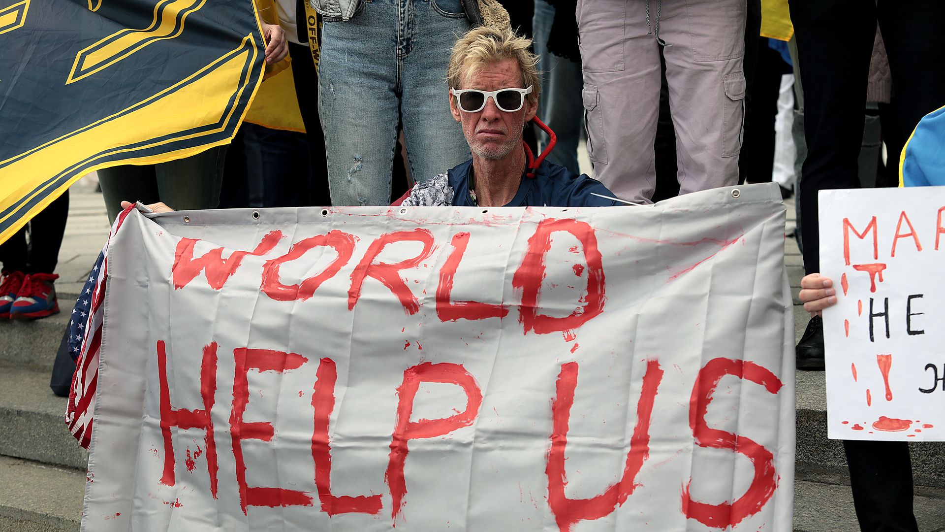 Ryan Routh, wearing white sunglasses, holds a white sign at a protest that reads "WORLD HELP US" in red letters.