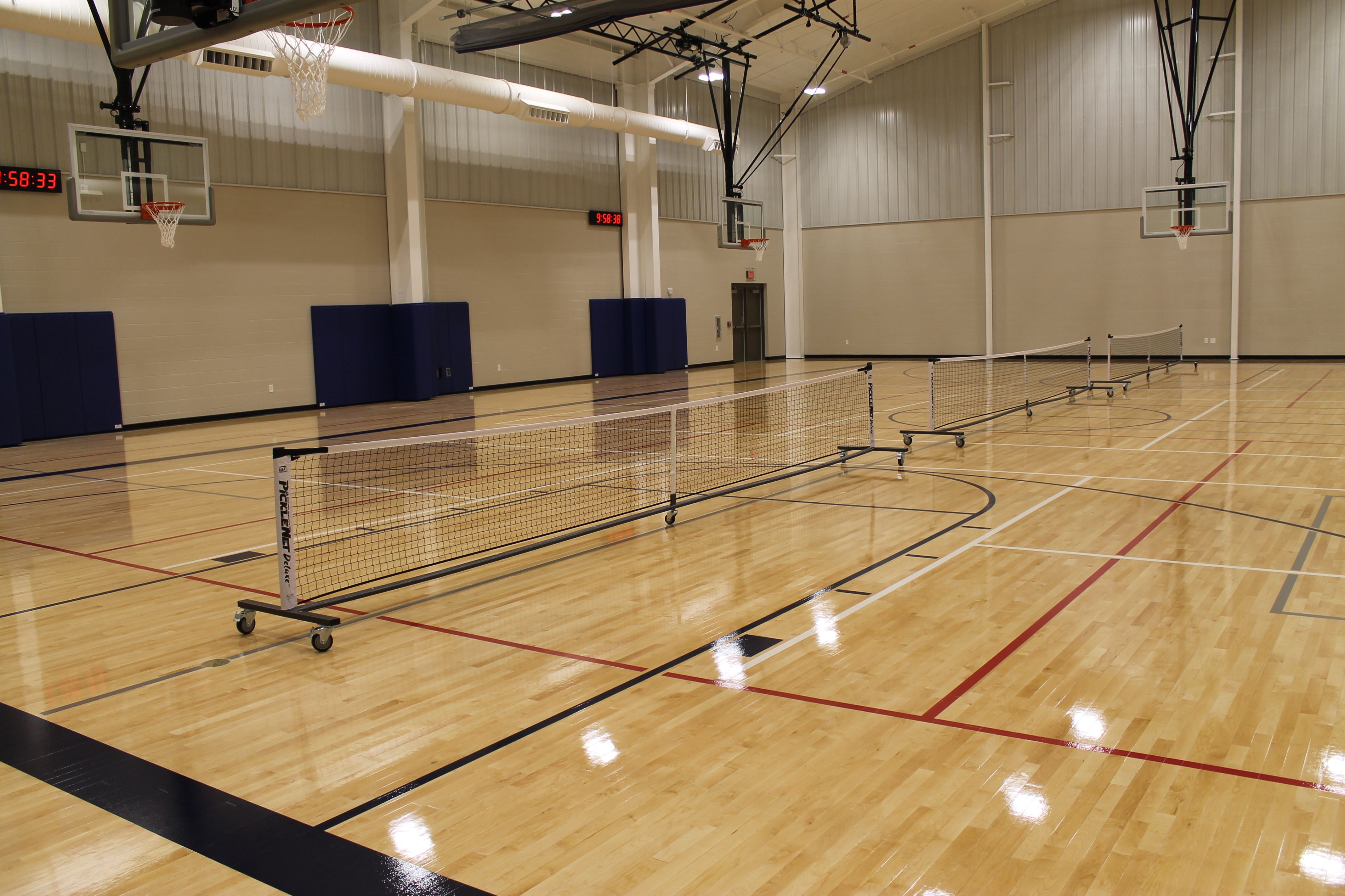 Indoor gymnasium with shiny wooden floor, multiple basketball hoops, and three portable pickleball nets set up across the court, walls are beige with blue padded sections.