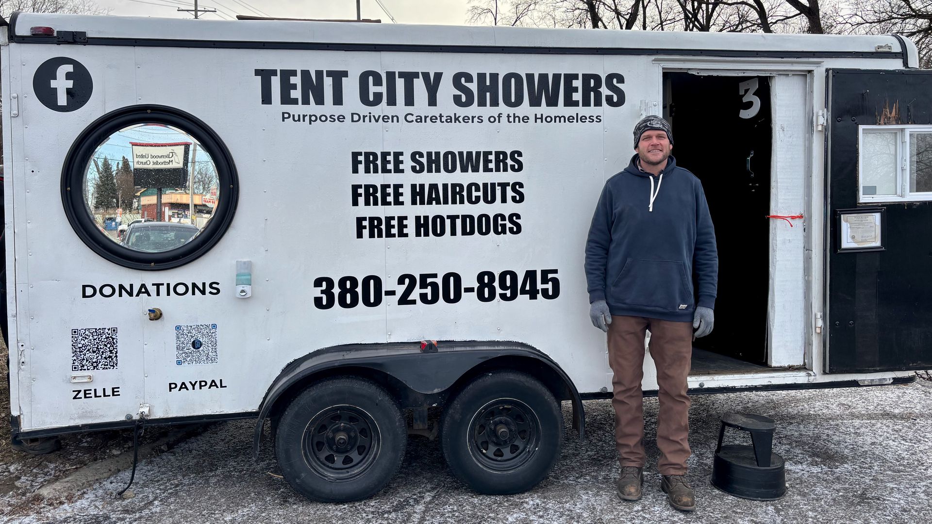 Man standing beside a white trailer labeled "Tent City Showers" offering free showers, haircuts, and hotdogs for the homeless, with donation QR codes and a phone number.