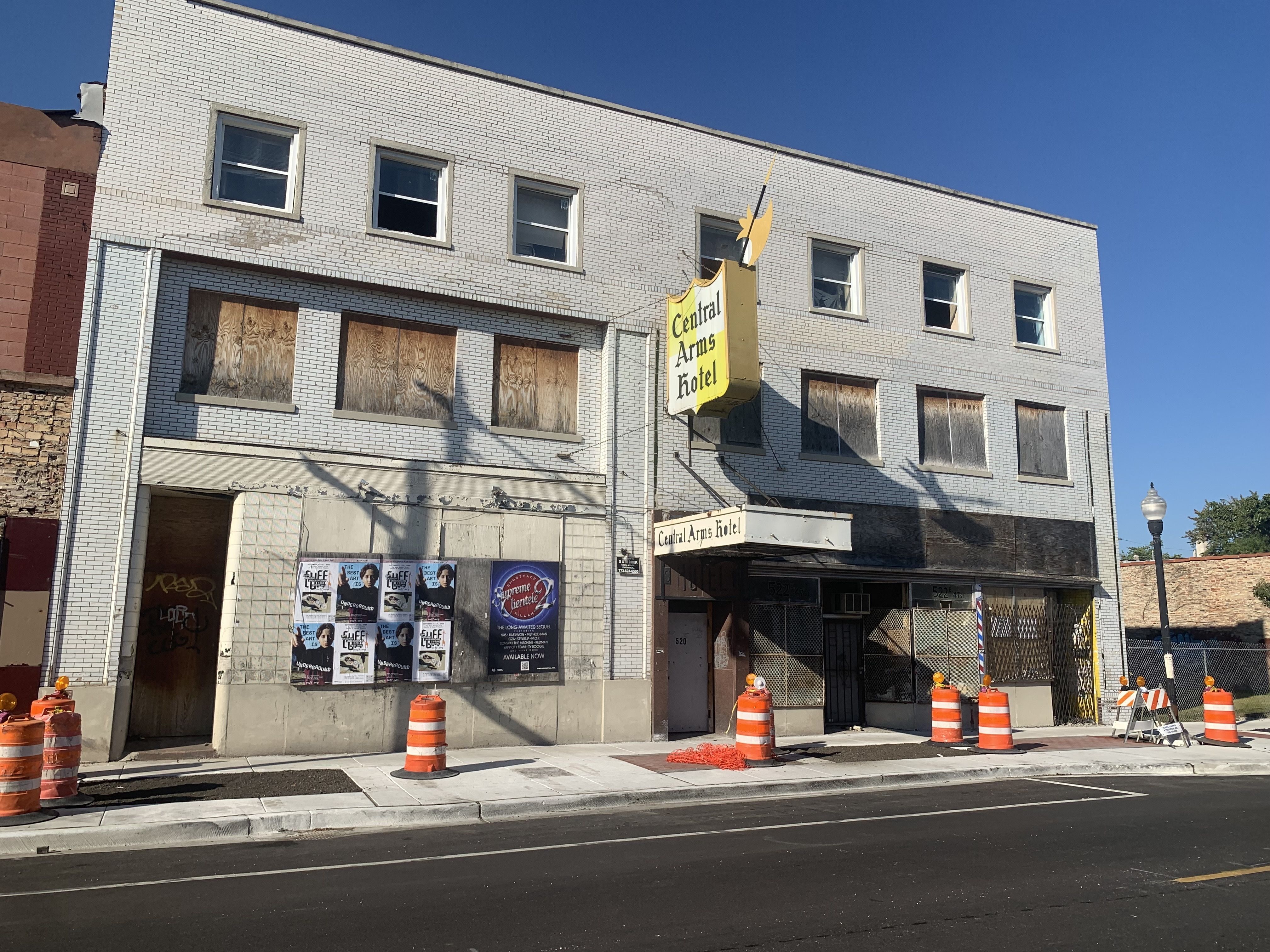 Old three-story white brick building with boarded windows and faded 'Central Arms Hotel' sign, surrounded by orange traffic barrels on a sunny day with clear blue sky.