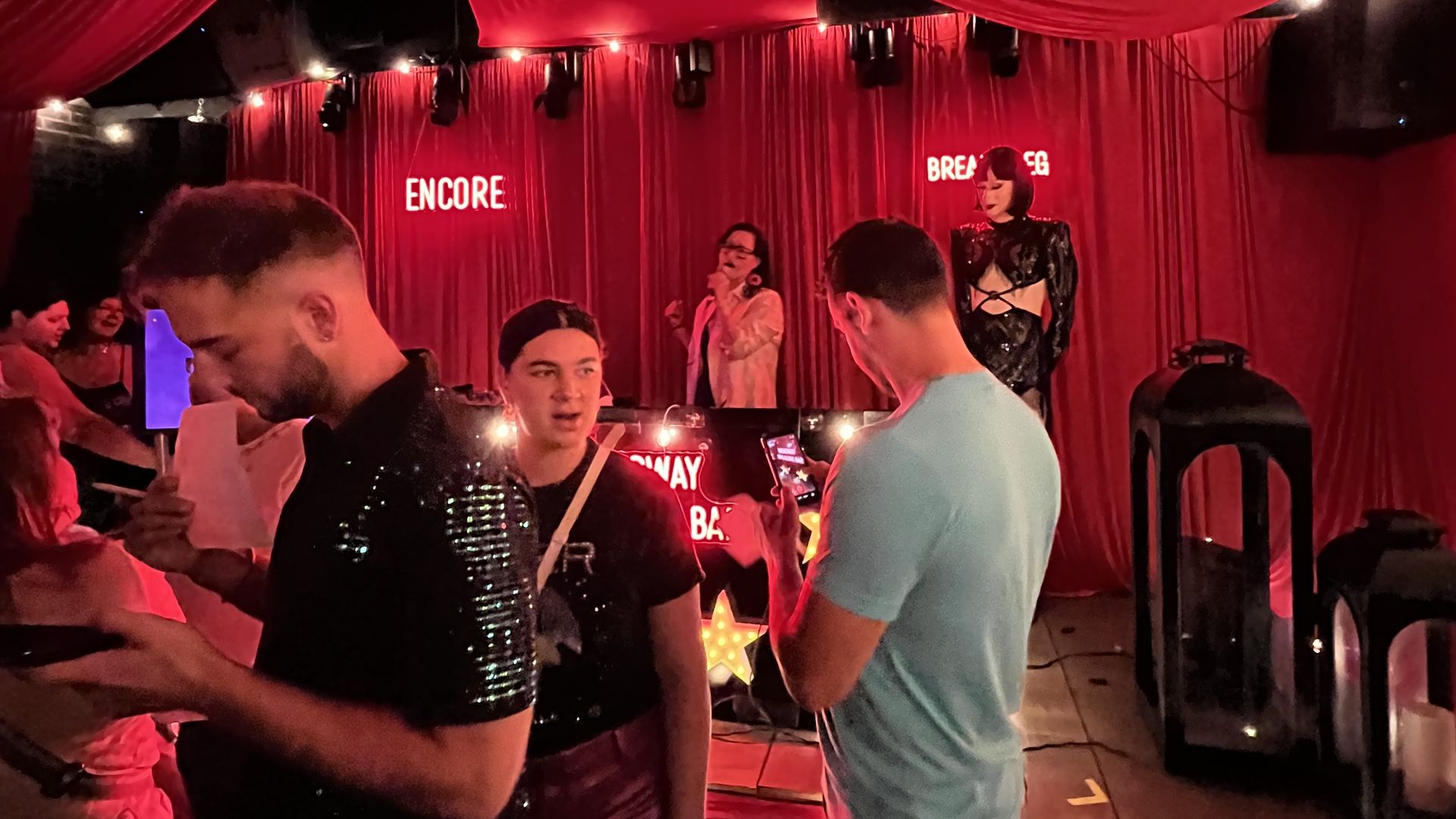 People socializing in a dimly lit room with red curtains, neon signs reading "ENCORE" and "BREAK" in the background, a performer in black lace, and large lanterns on the floor.