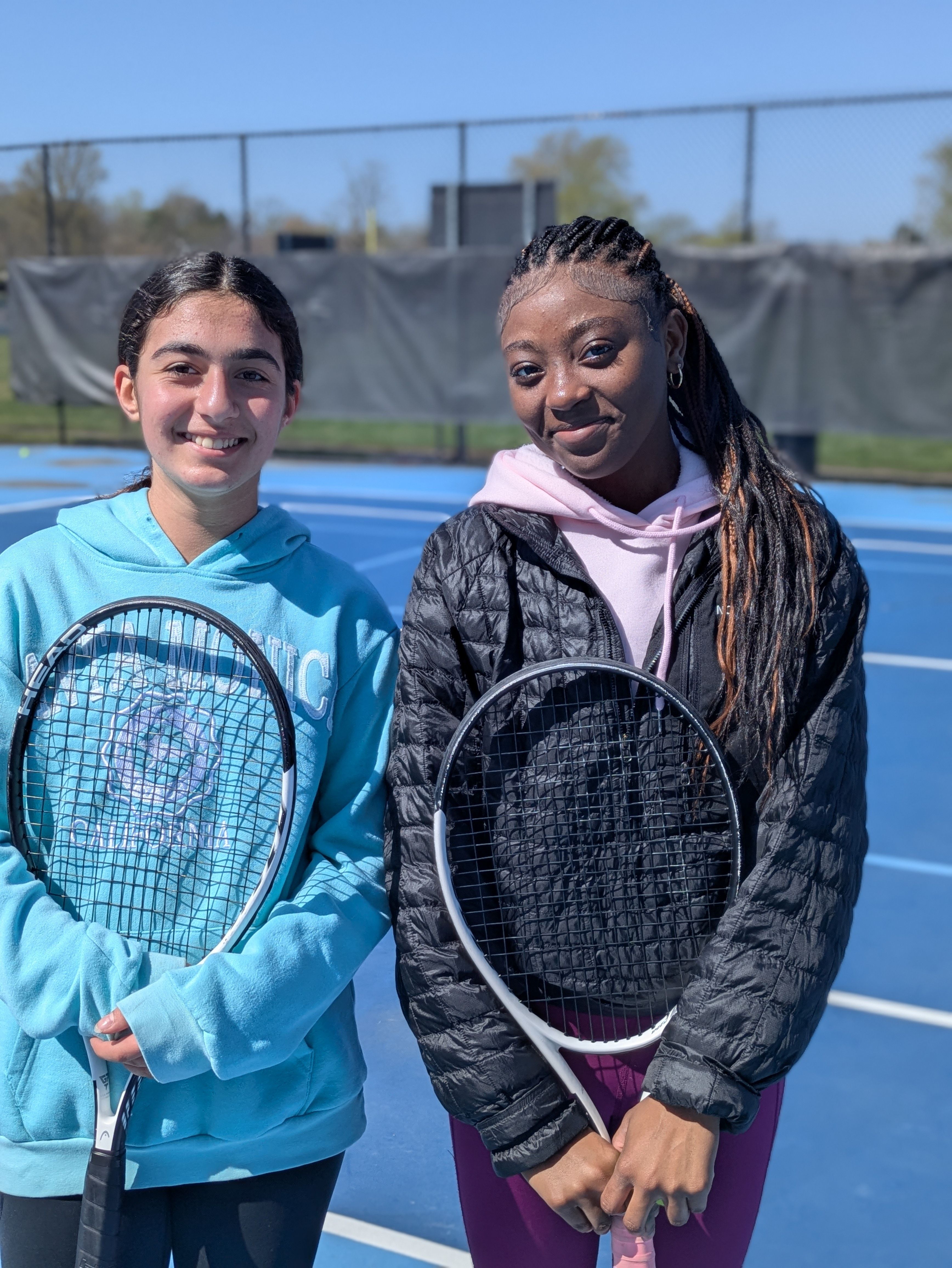 Two young women stand on a blue tennis court, each holding a racket. Left wears a light blue hoodie; right, a black quilted jacket over pink hoodie and purple leggings. Both smile at camera.