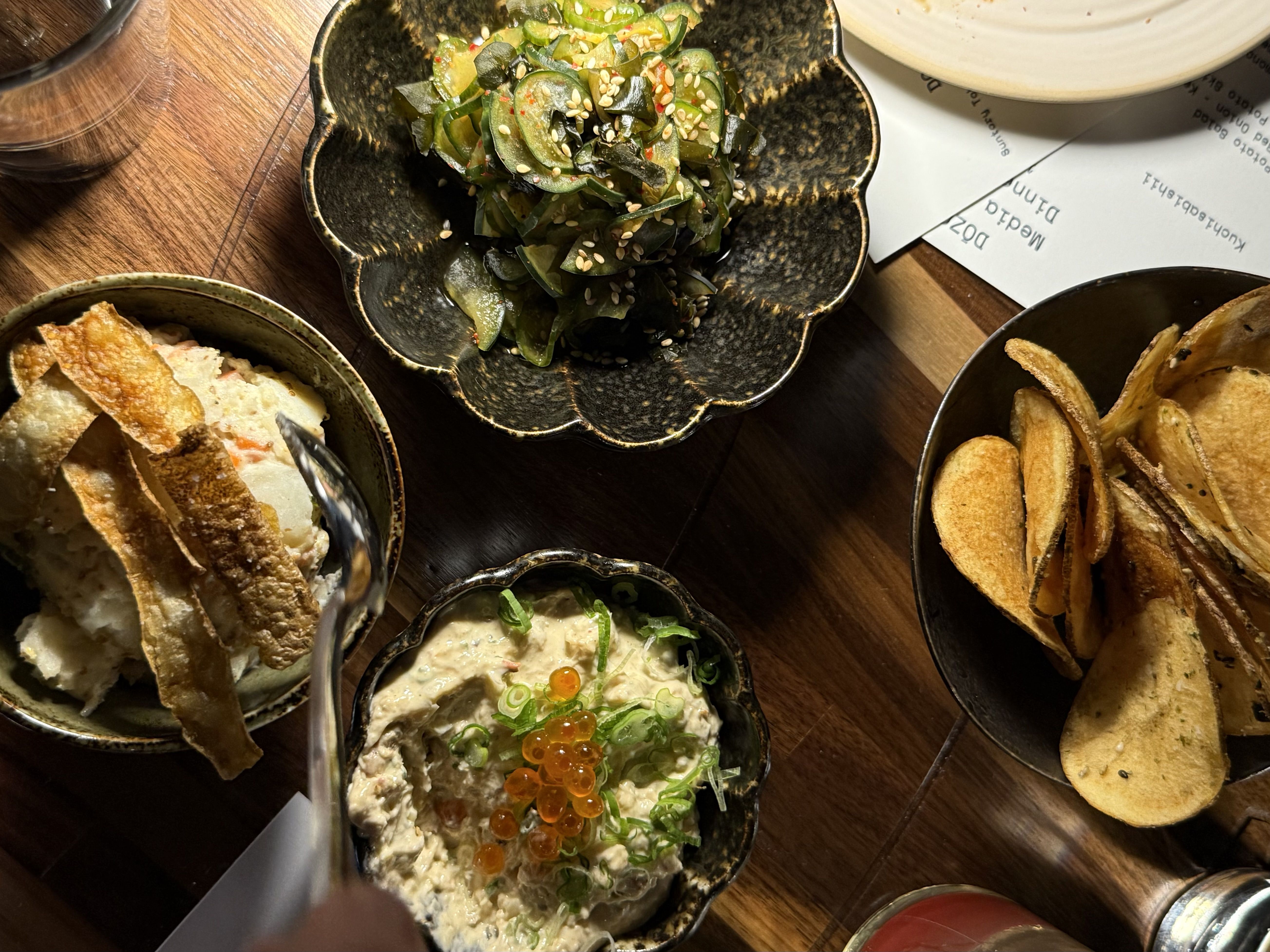 Overhead view of various dishes on a wooden table including potato salad with crispy skin, seaweed salad sprinkled with sesame seeds, a creamy dip topped with green onions and orange roe, and a bowl of potato chips.