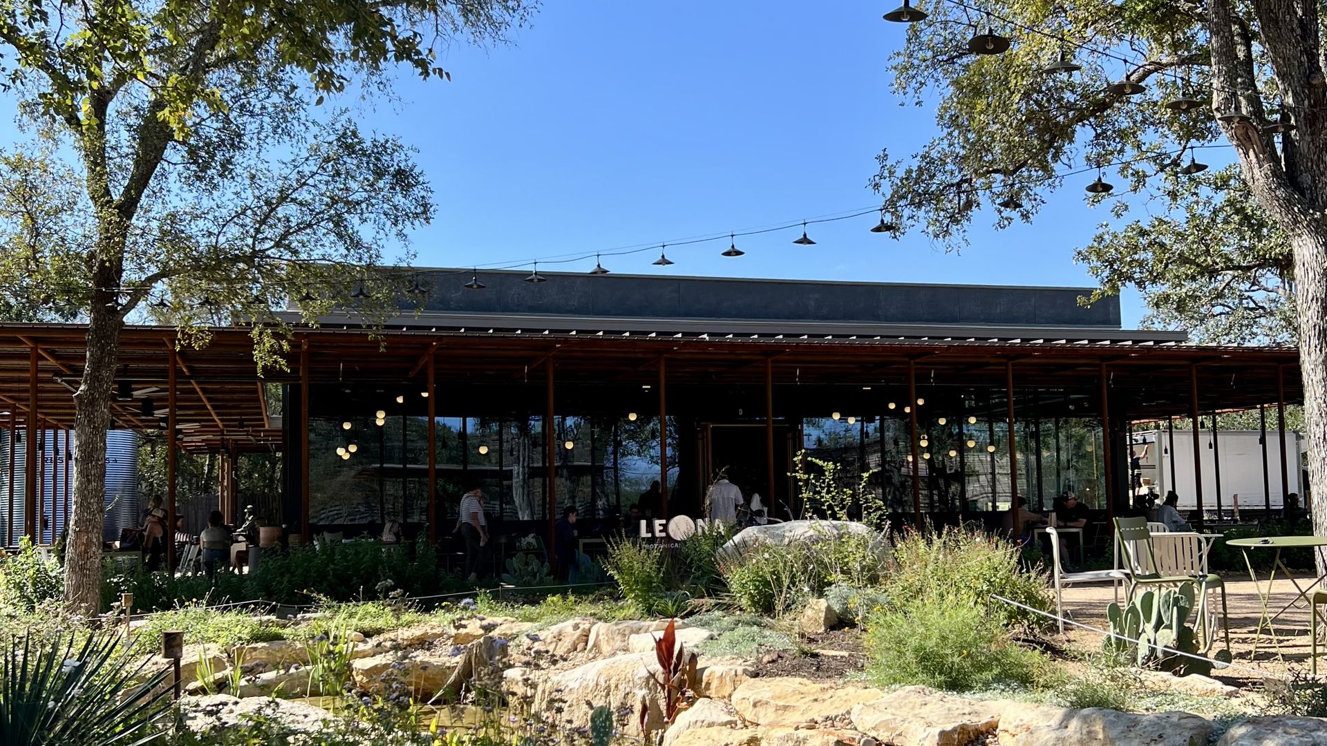 Outdoor café scene with wooden patio and string lights hanging between trees under bright blue sky, surrounded by green plants, rocks, and customers sitting at tables.