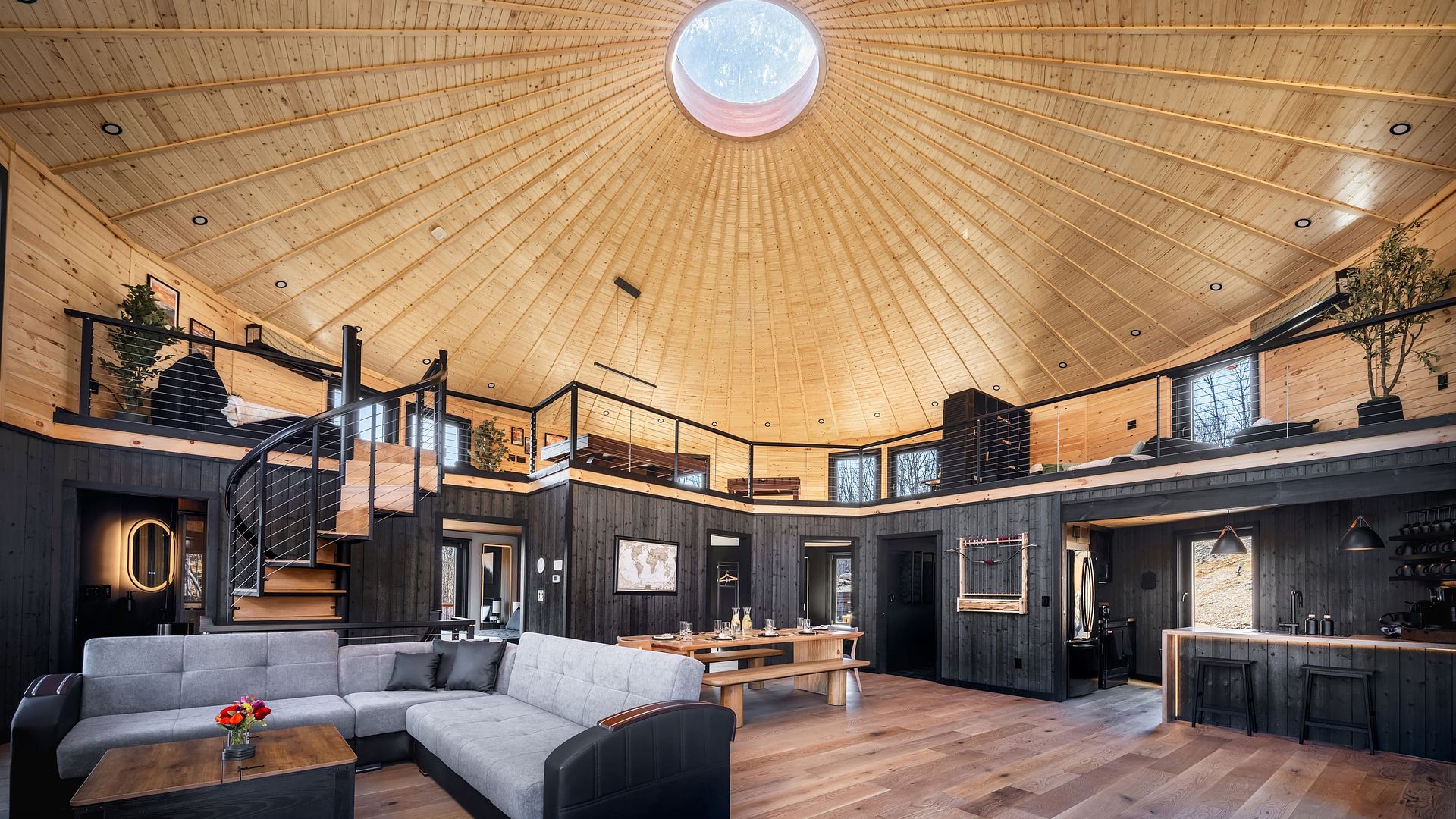 Interior of a circular, wood-paneled room with a domed ceiling and a round skylight. A black-railed mezzanine overlooks a modern living space with a gray sectional, wooden tables, and a kitchen.