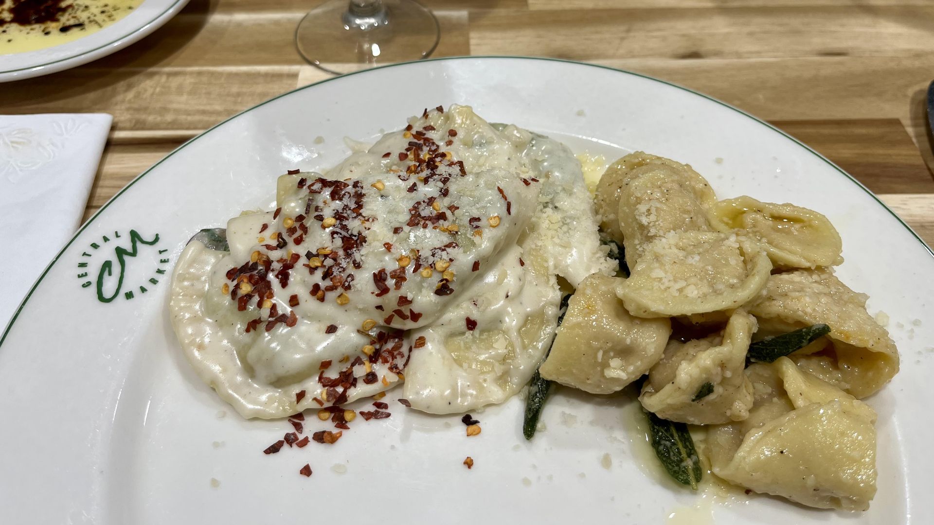 A white Central Market plate with a green rim holds two homemade pastas: one is a creamy ravioli topped with red pepper flakes, the other is a tortellini with butter and sage. A wine glass and a water class sit on a wooden table in the background.