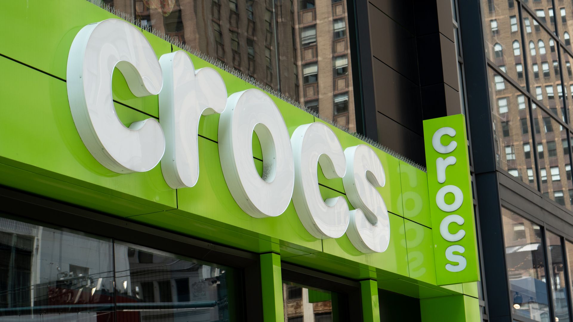 Close-up of a bright lime-green storefront with large white 3D letters along the facade; a row of spikes along the top edge. Glass reflects tall brick buildings on a city street.