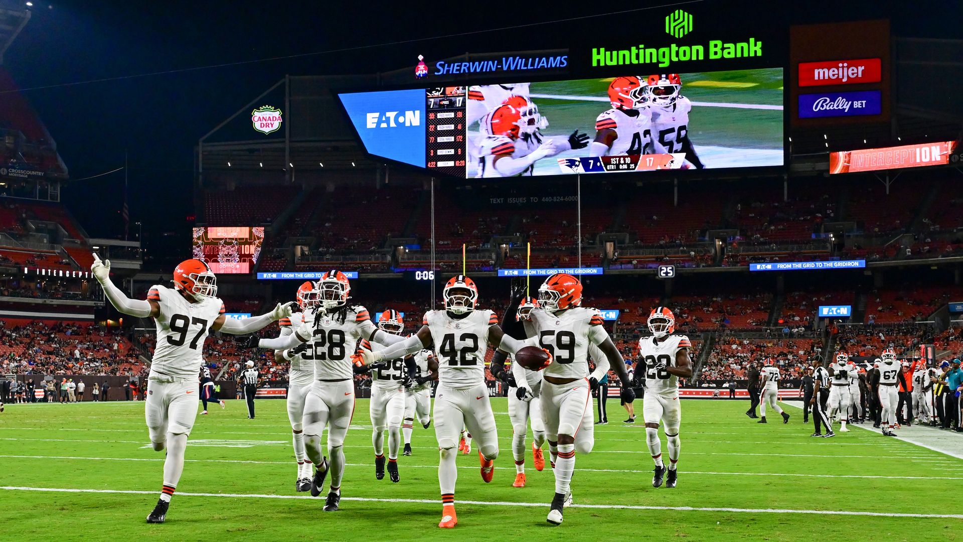 Night football game. White-uniform players with orange helmets celebrate on the field as a big scoreboard shows game action and sponsor logos like Eaton, Sherwin-Williams, and Huntington Bank.
