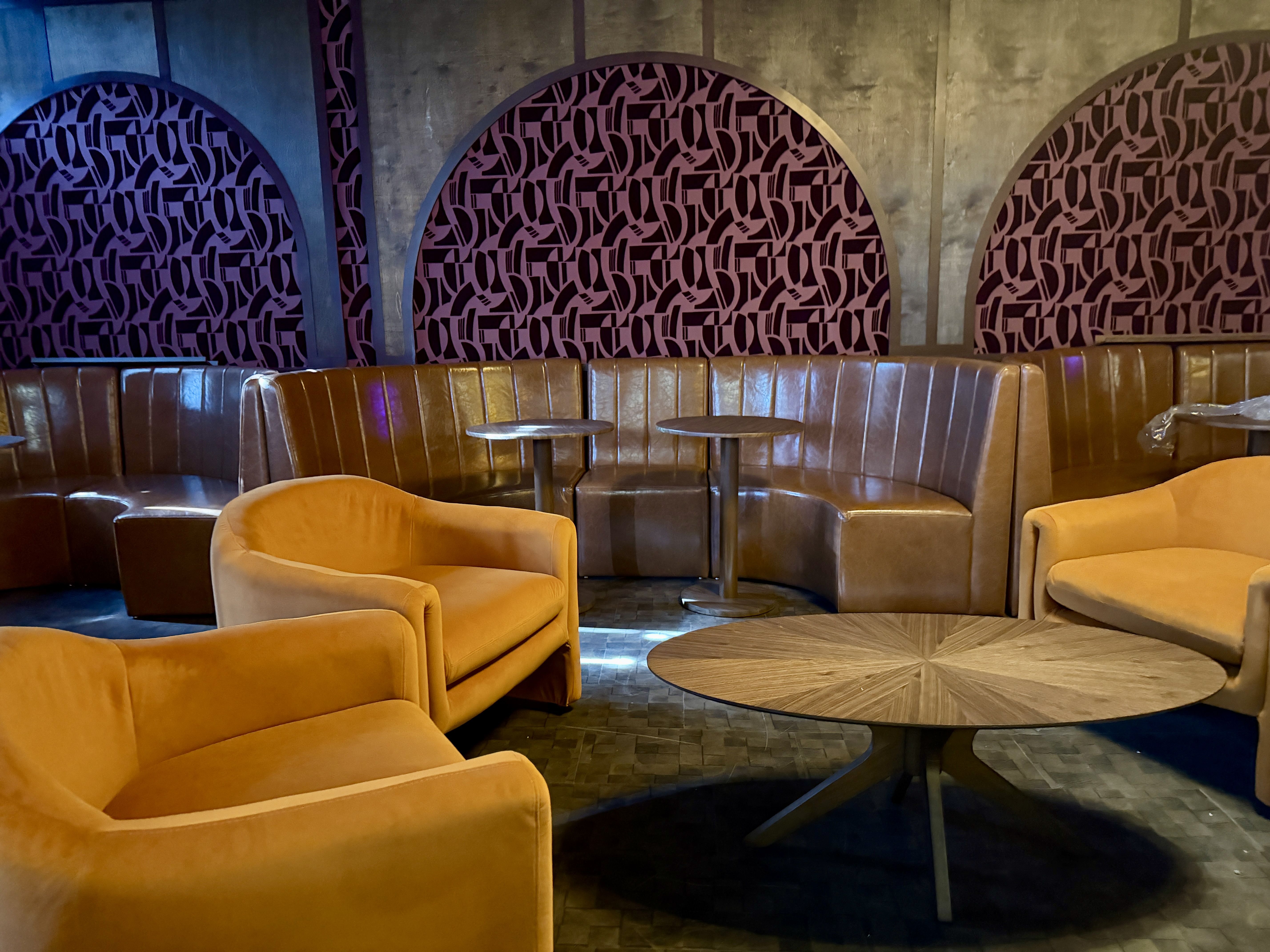 Cozy lounge with mustard yellow armchairs, brown leather curved bench seating, round wooden tables, and decorative wall panels with purple and black geometric patterns.