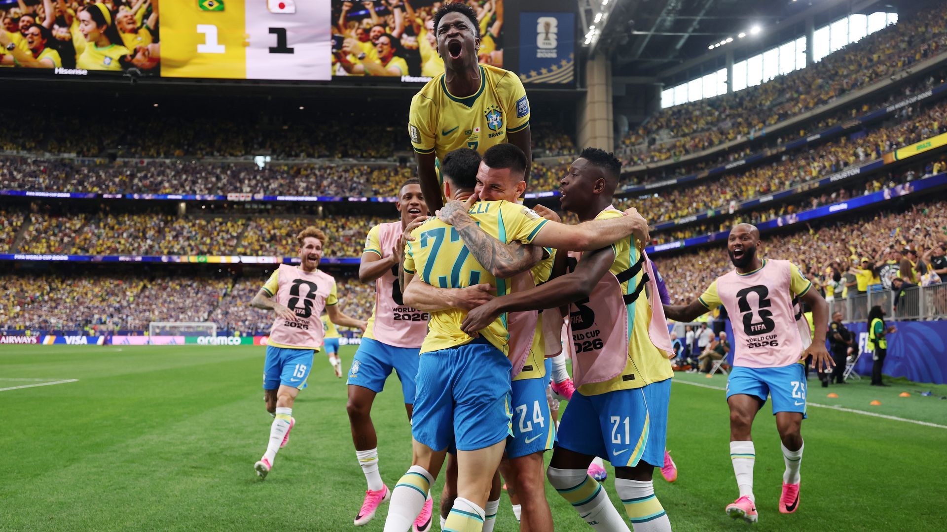 Brazilian soccer players in yellow jerseys celebrate a goal, lifting a teammate as others hug, in a packed stadium with yellow-clad fans and pink training bibs.