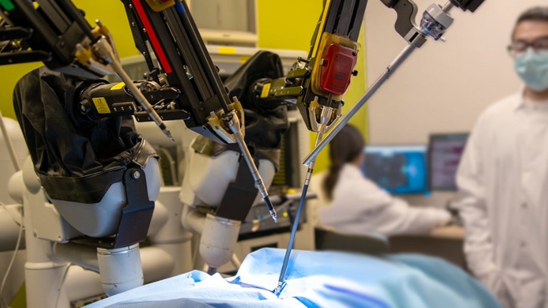 Robotic surgical system with several articulated arms performing minimally invasive surgery on a patient draped in blue, as clinicians in white coats and masks monitor in the background.