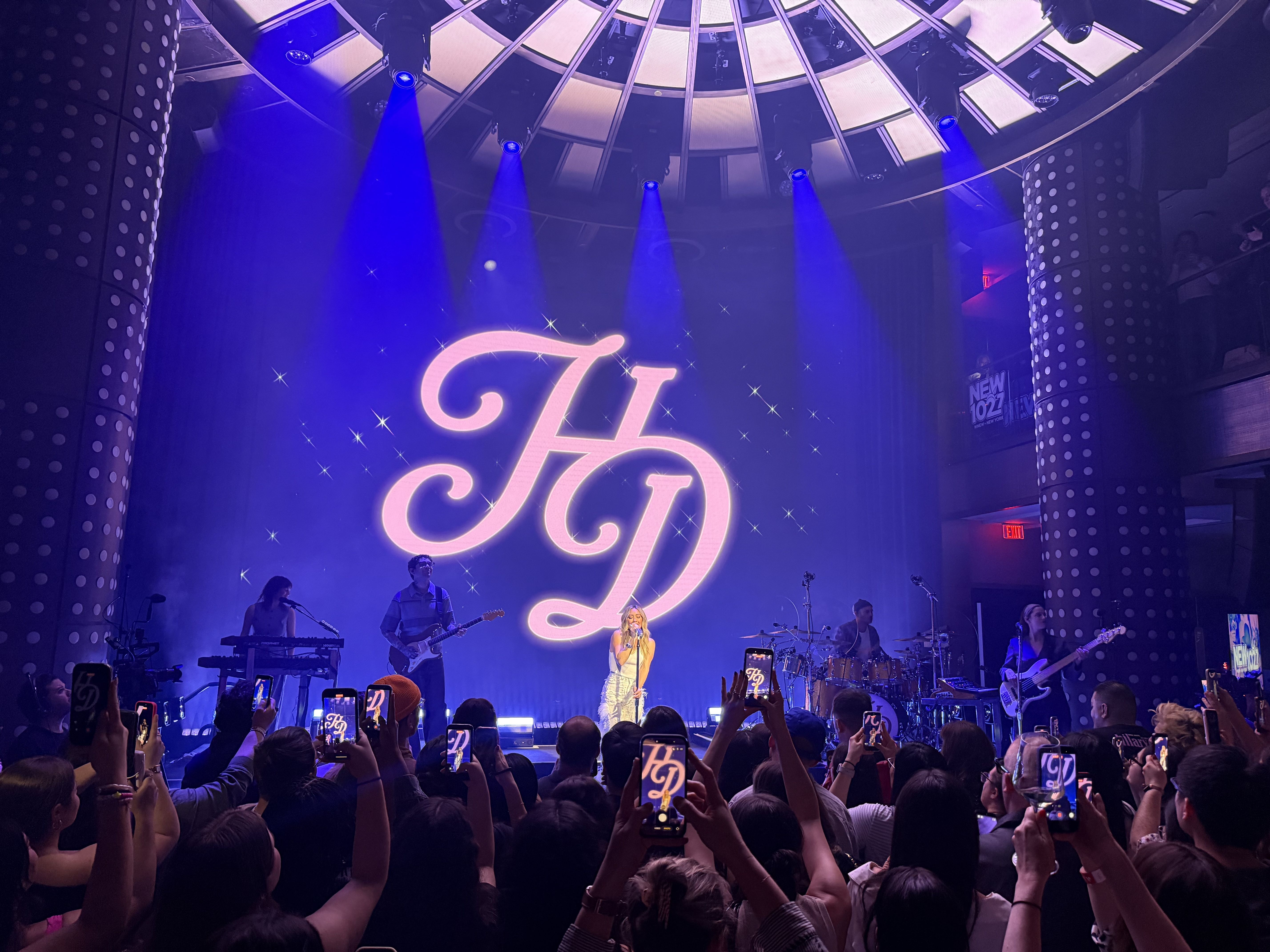 Concert stage with a neon pink HD logo backdrop, blue spotlights, and a starry curtain. A band performs with a female singer center stage while audience in the foreground holds up smartphones.