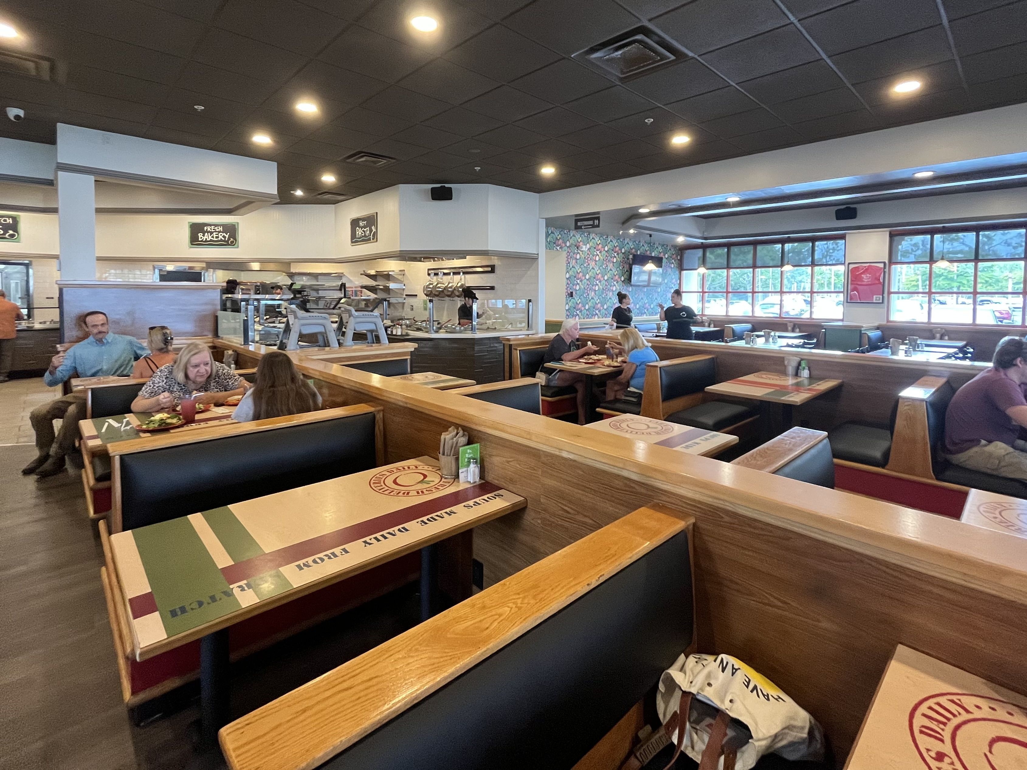 Casual restaurant with wooden booths and long tables. Open kitchen in back; dark ceiling with recessed lights. Diners sit by large windows; colorful wallpaper on the back wall; signs read "Fresh Bakery" and "Hot Pasta".