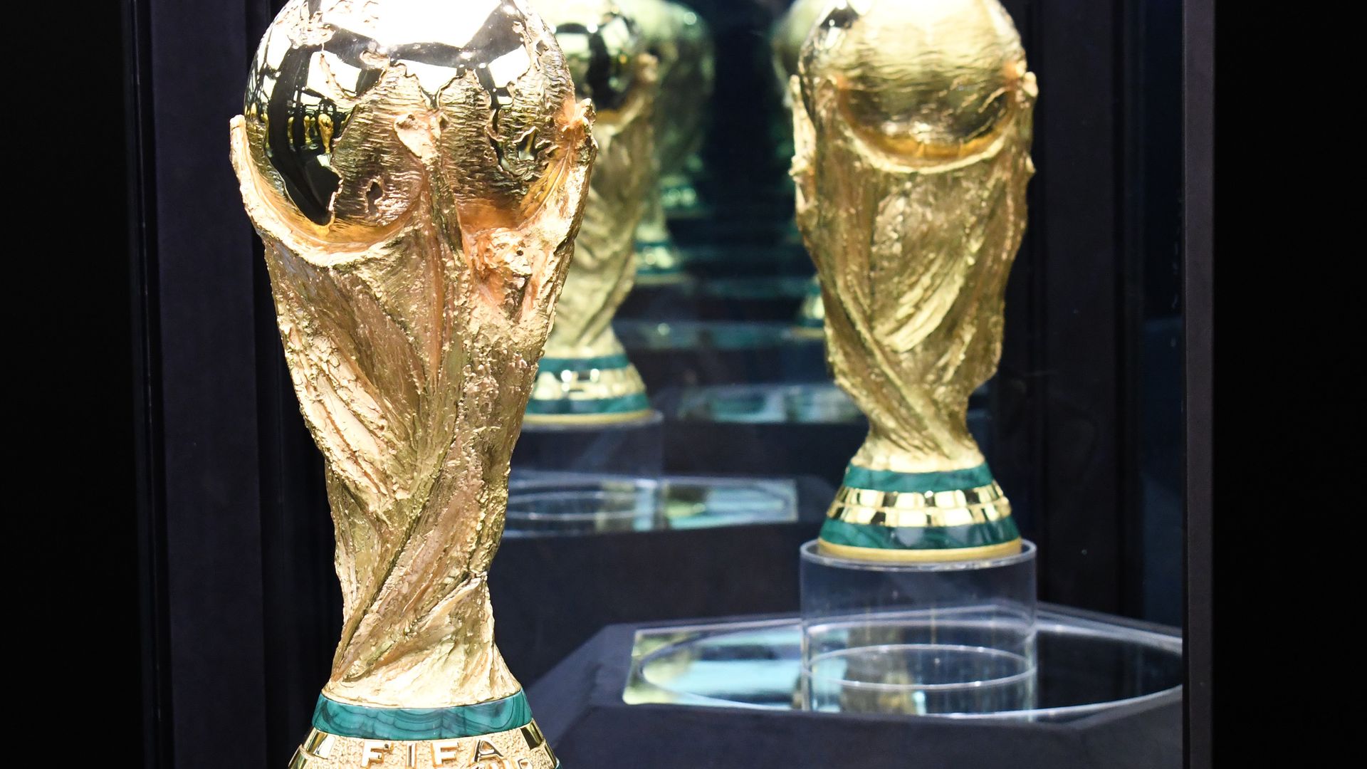 A golden trophy with a green base inscribed with "FIFA WORLD CUP" sits on a pedestal and is reflected in a nearby mirror. 