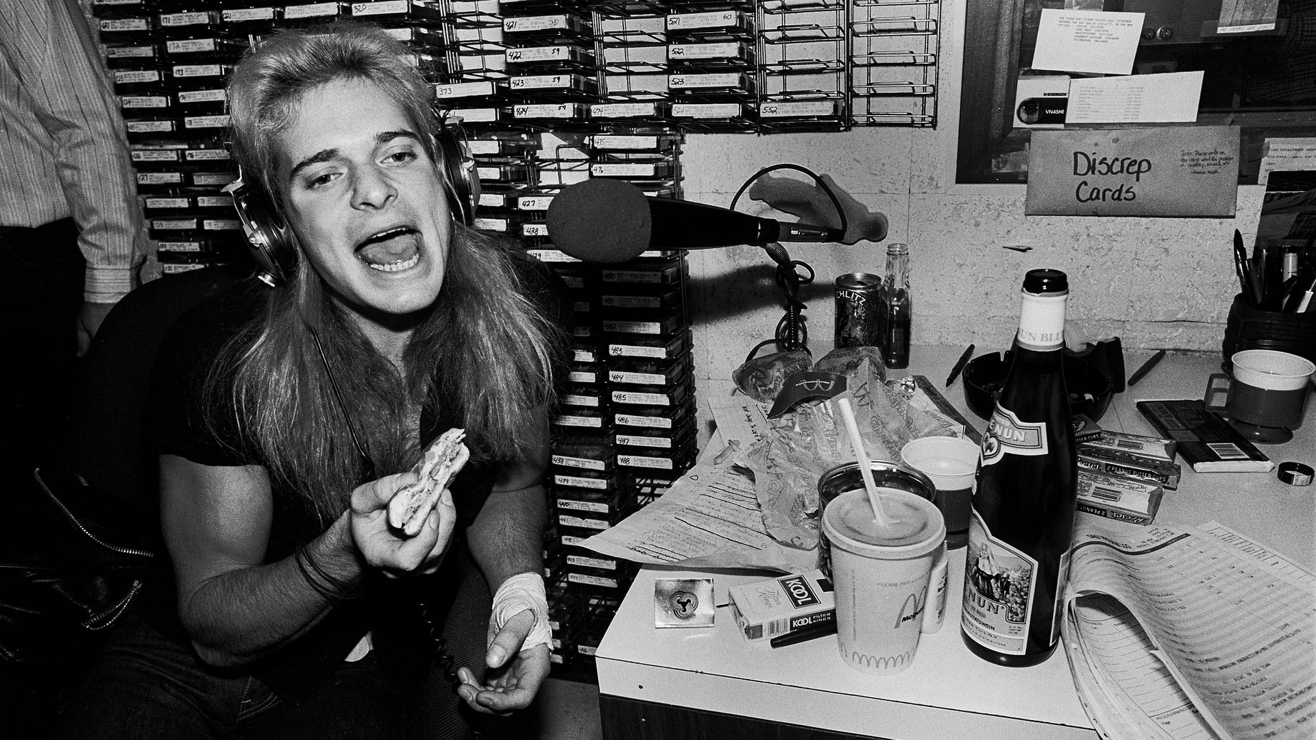 Photo of a man talking into a microphone while eating a sandwich. 