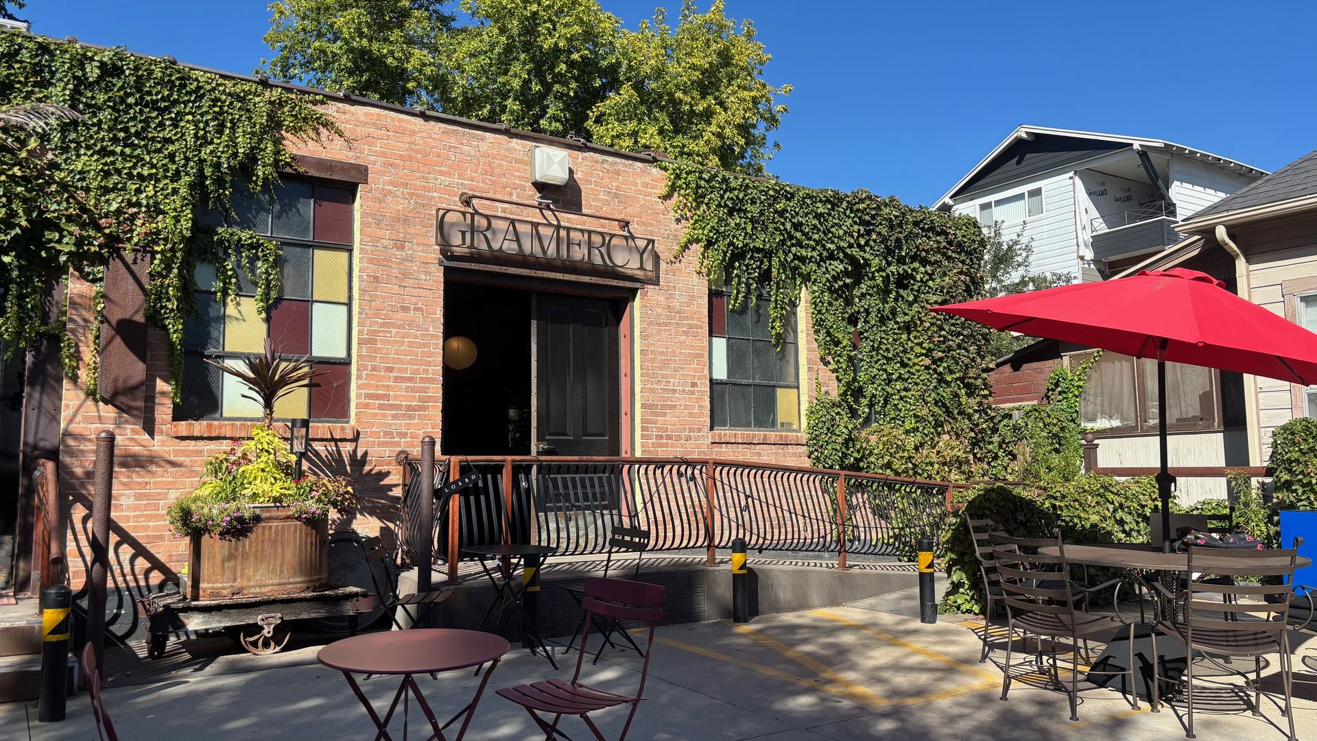A brick building covered in ivy with a metal sign reading "GRAMERCY" above the entrance. Outdoor seating with red umbrella, metal chairs and tables, and a clear blue sky.