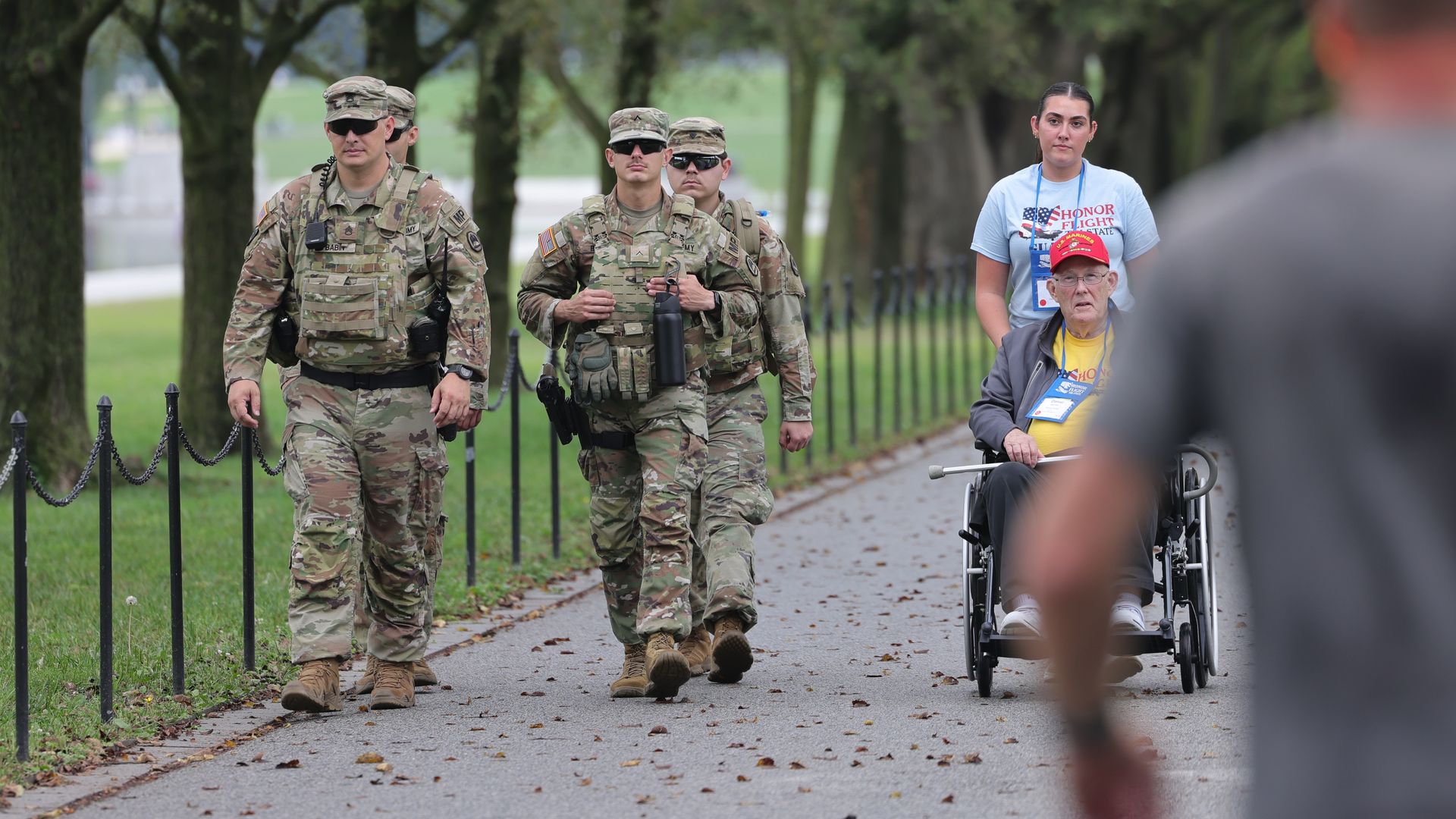 Four soldiers in camouflage uniforms walking on a tree-lined path, while a woman in an "Honor Flight" shirt pushes an elderly man in a wheelchair wearing a red cap. 