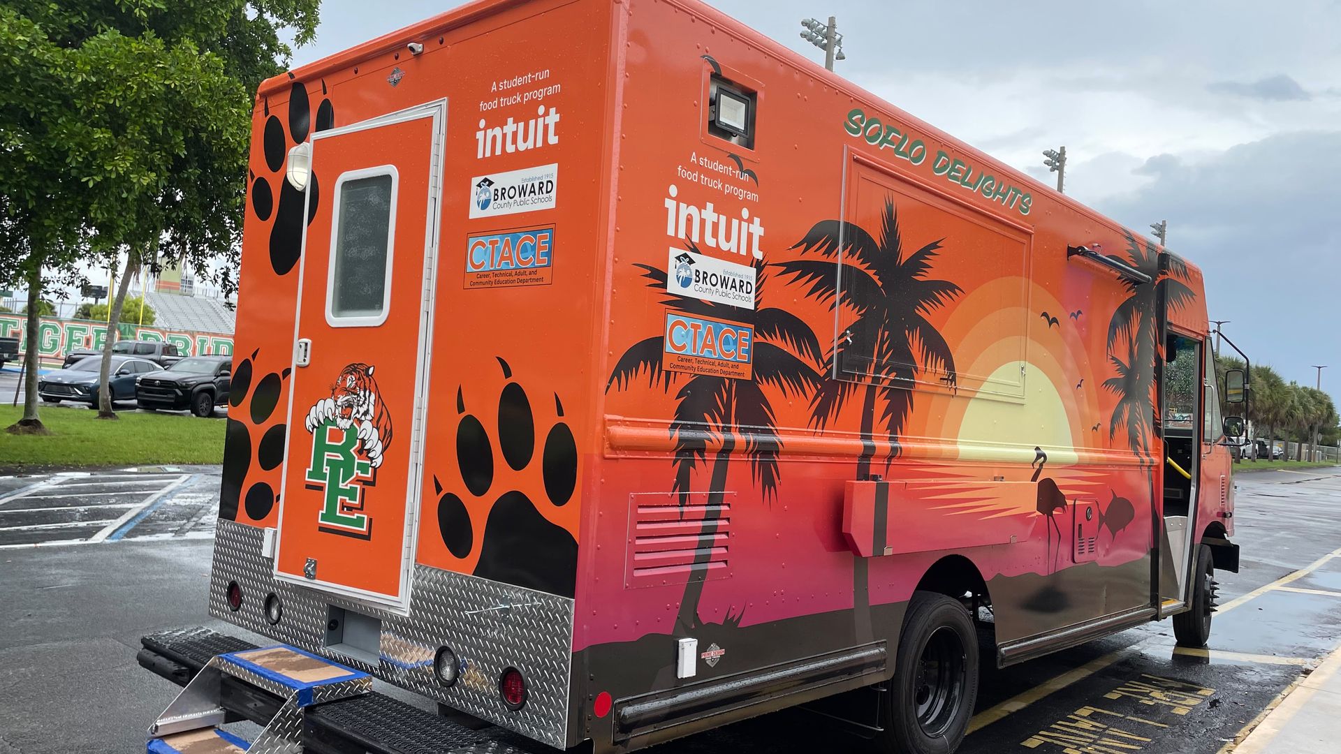 Bright orange food truck with palm trees and the word Intuit on its side, "SOFLO Delights" along the top, plus sponsor logos. A green tree and cloudy sky above a parking lot.