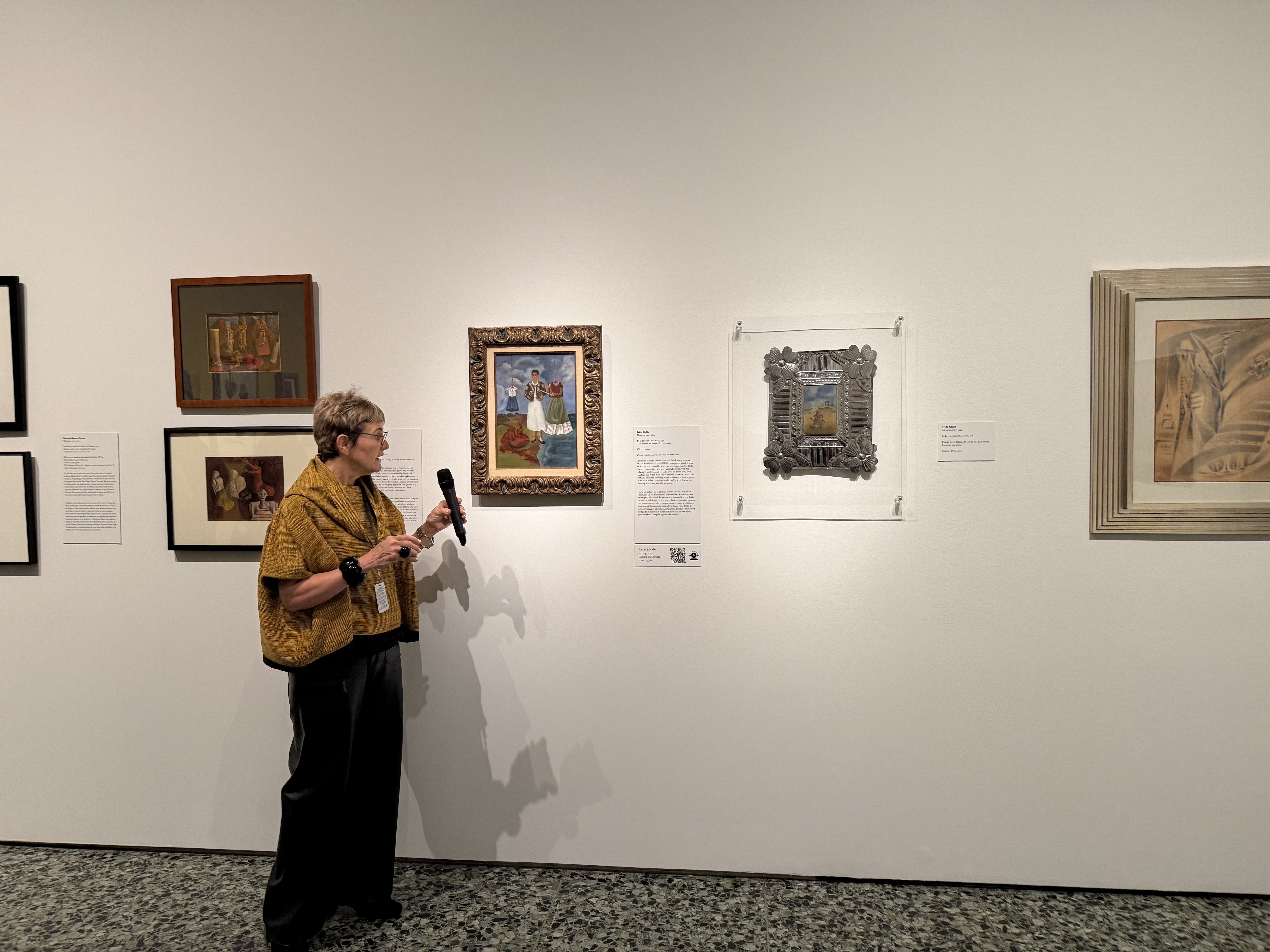 A woman in a mustard shawl and dark pants holds a microphone while standing near framed paintings on a beige gallery wall with informational plaques.