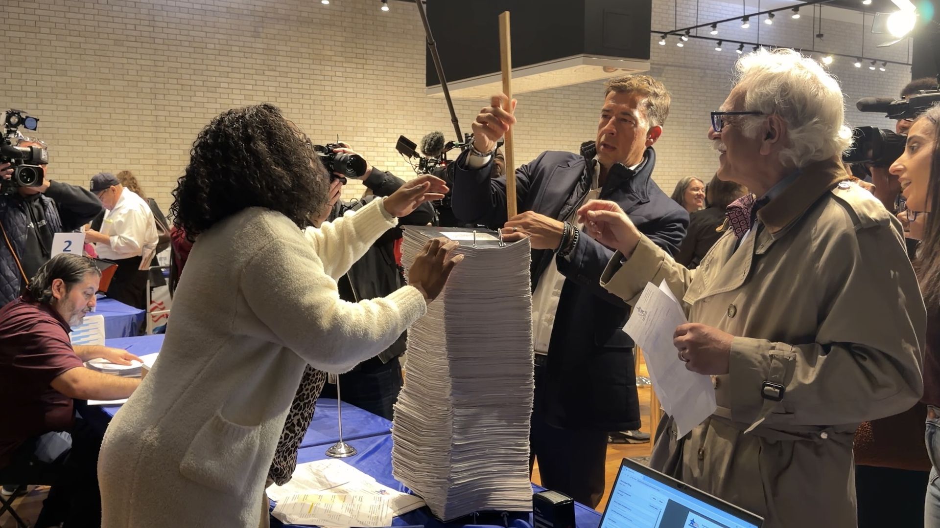News reporters and bystanders surround a blue table with a tall stack of papers as a man raises a wooden rod over it; cameras film in a bright brick-walled room.