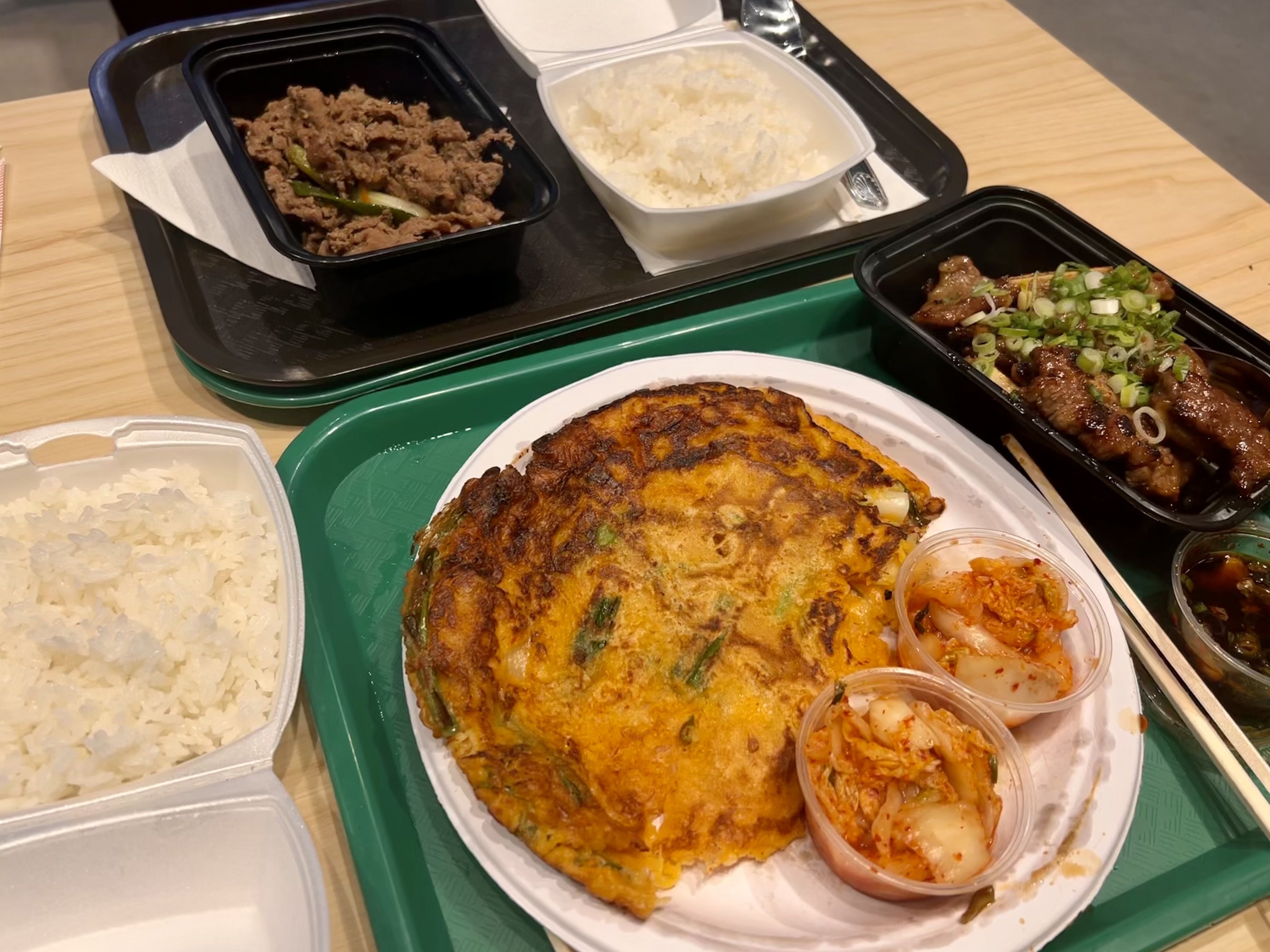 A picture of rice, a kimchi pancake with two sides of kimchi, beef bulgogi in a black container and ribs in another black container on trays.