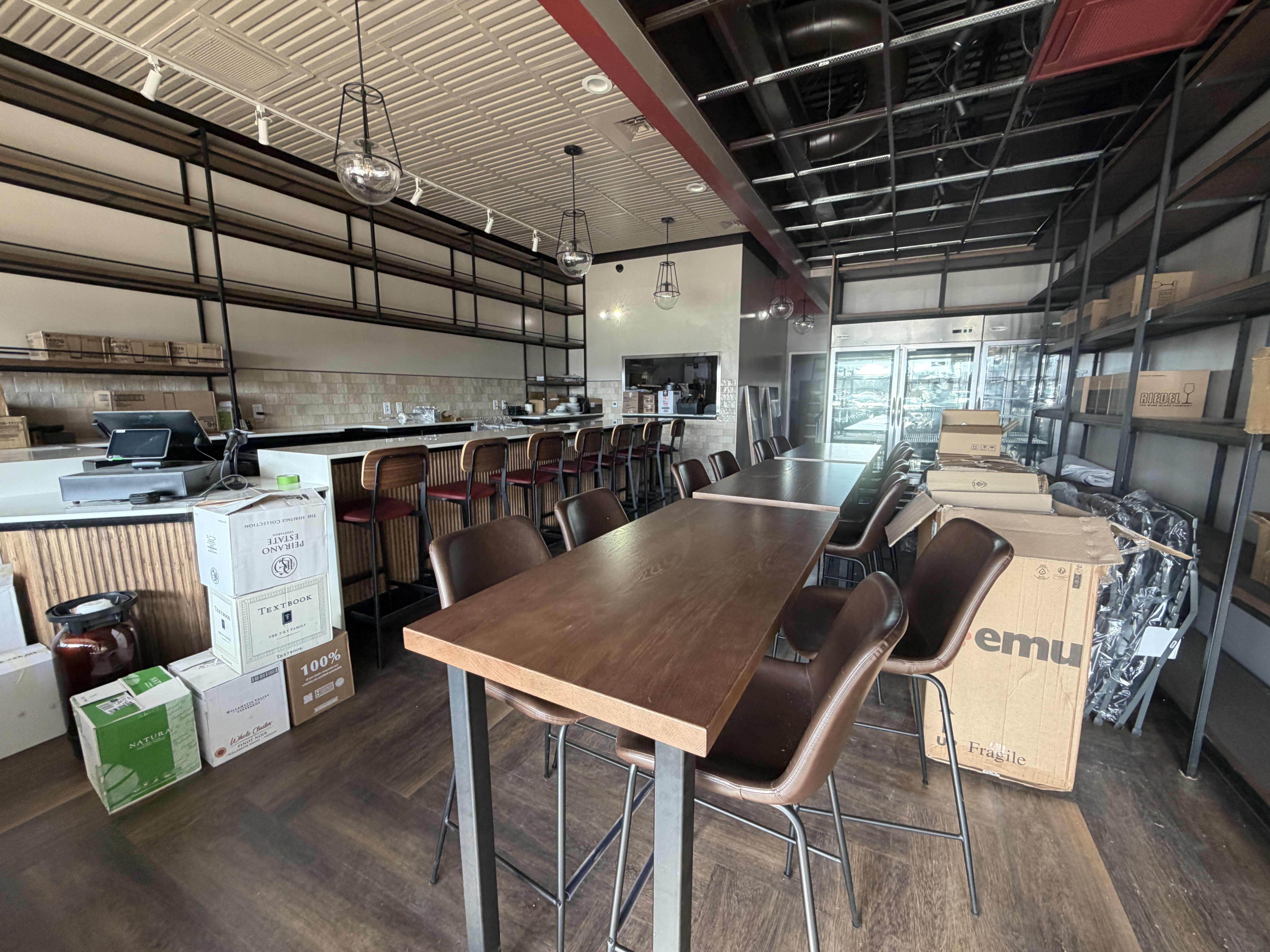 Indoor bar area with wooden tables, brown chairs, bar stools with maroon seats, modern lights, and shelves. Cardboard boxes and packaging materials scattered around, indicating setup or storage.
