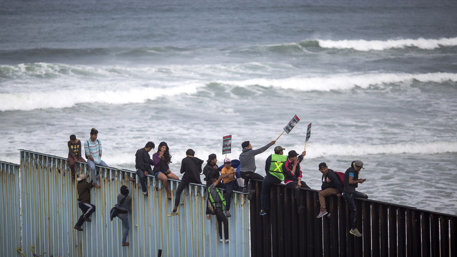 People climb a section of border fence to look toward supporters in the U.S.