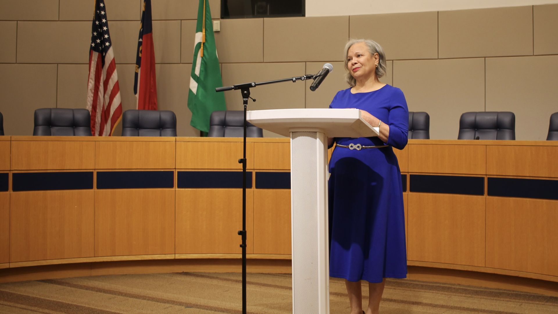A woman in a royal blue dress stands at a white podium with a microphone in a formal conference room; three flags, including the U.S. flag, a state flag, and a green flag, line the tan wall behind her.